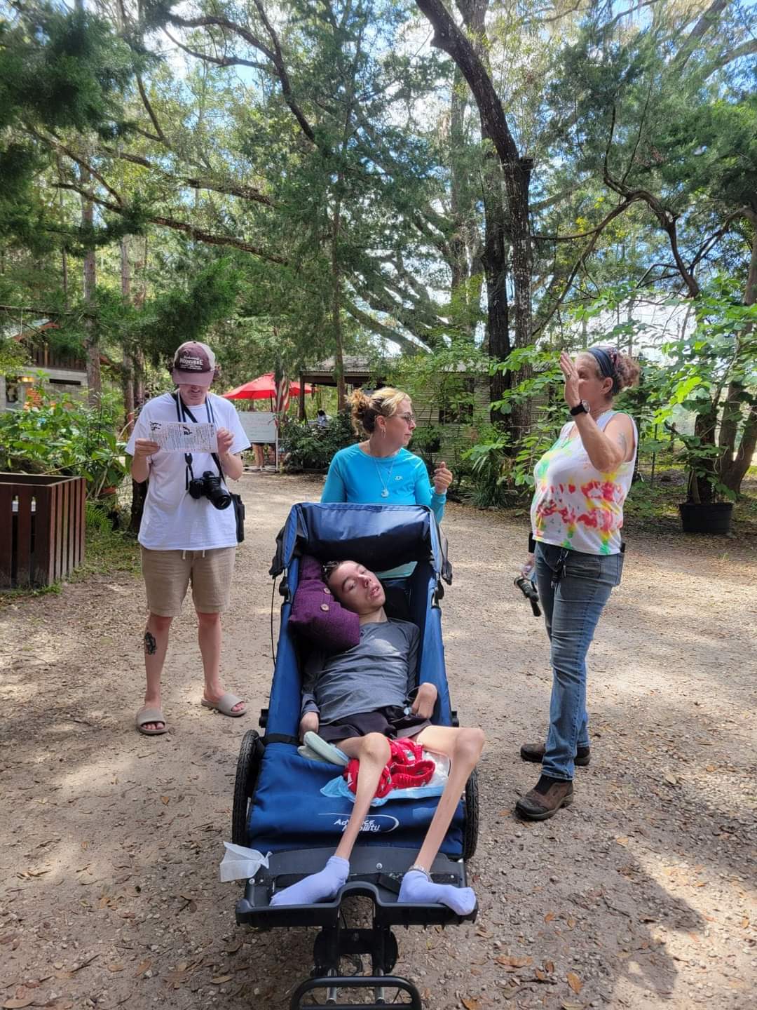 Four people outdoors in a park, two women standing and talking, one woman with a camera, one person sitting in a stroller, trees in background