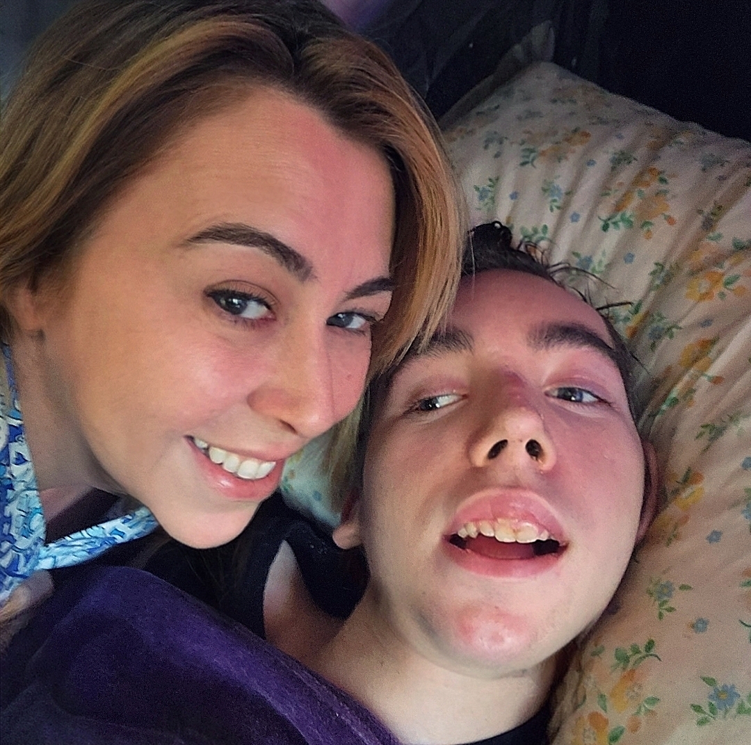 Two people lying on a bed, smiling at the camera, with a floral pillow in the background.