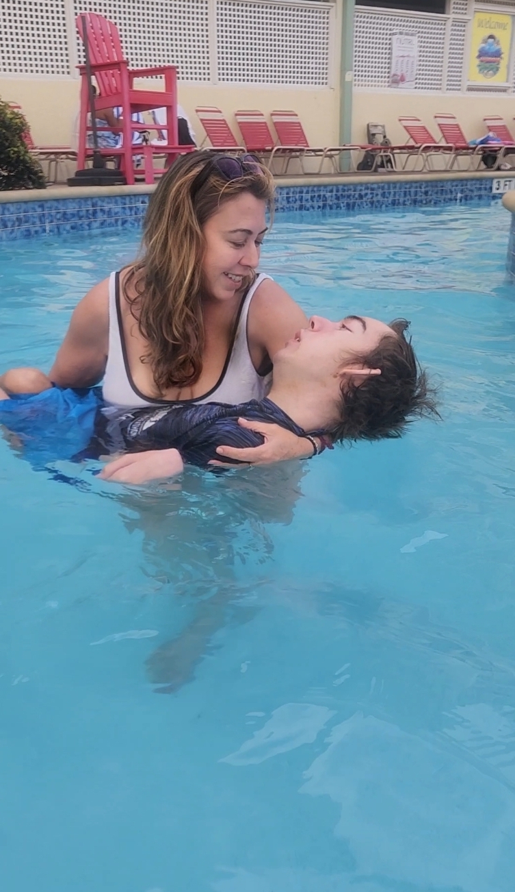 Two women in a swimming pool, one holding the other, smiling and looking at each other.