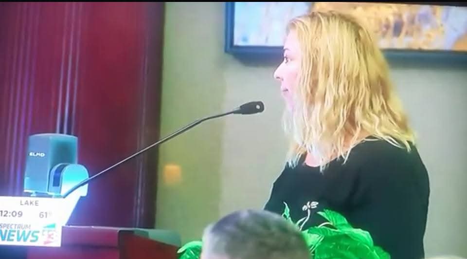 Woman with blonde hair speaking into a microphone at a podium in a room with green walls and a framed picture.