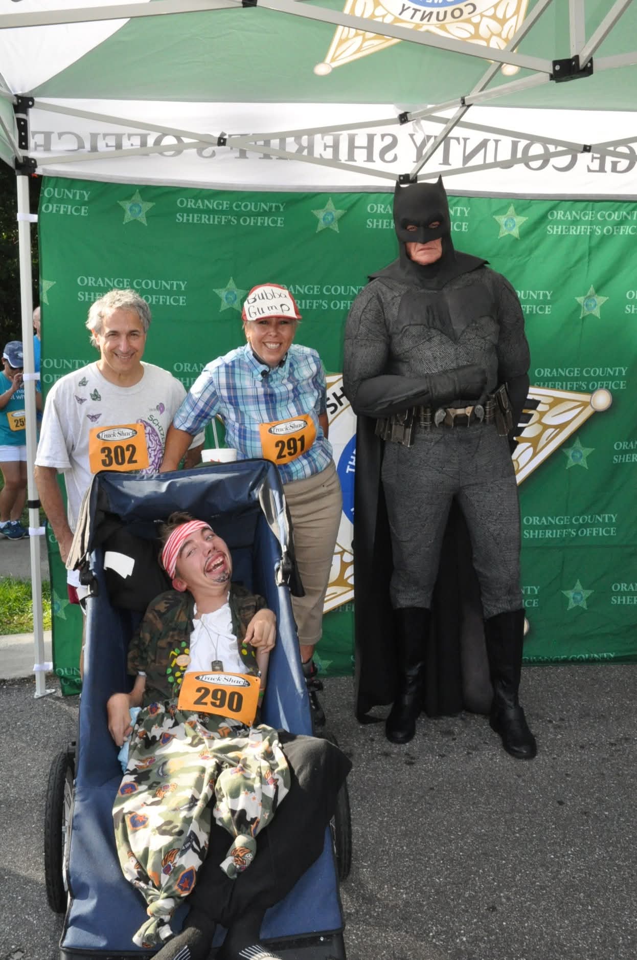 Three people and a person dressed as Batman at an outdoor event, with a green backdrop and a canopy overhead.