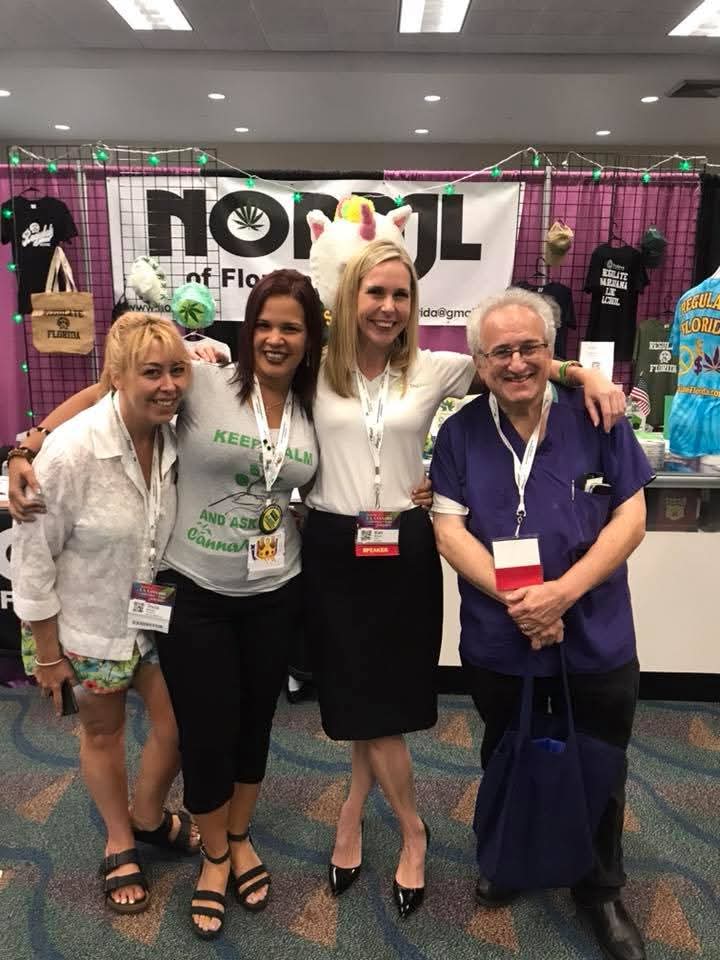 Four people standing together at an event, smiling, with a colorful booth in the background, including a banner and various items.
