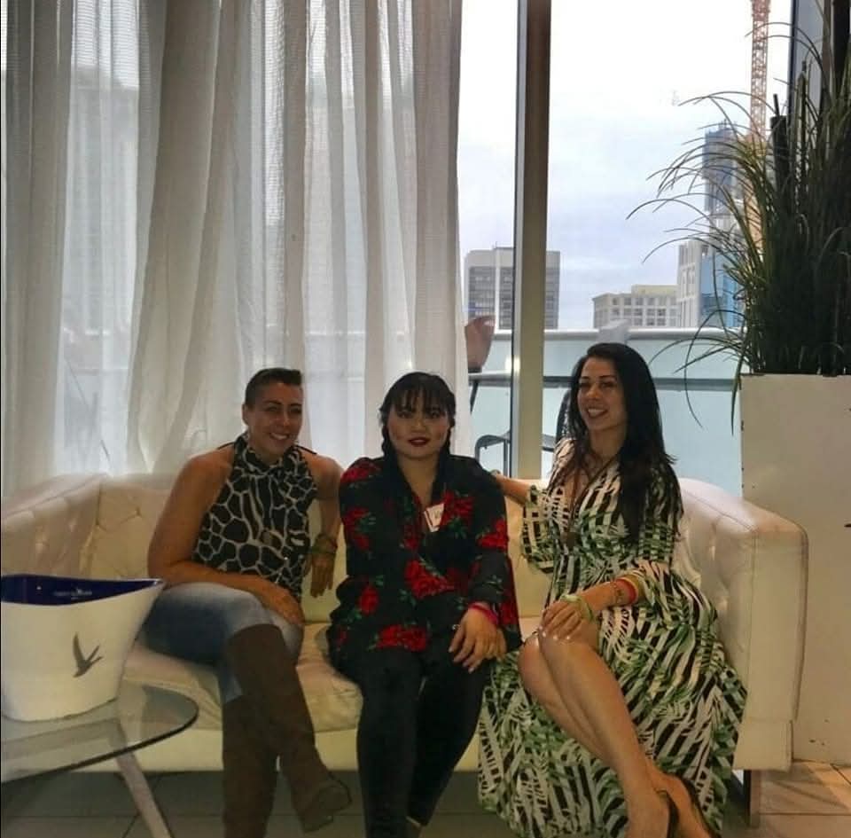Three women sitting on a white sofa near a large window with curtains, with buildings visible outside, and a tall plant nearby.
