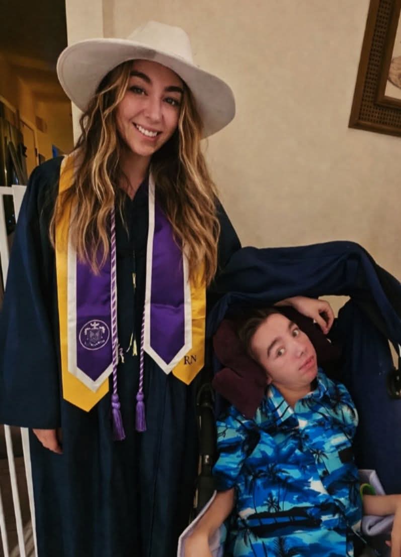 Young woman in graduation gown and hat smiling, standing next to a person in a blue patterned shirt, indoors.