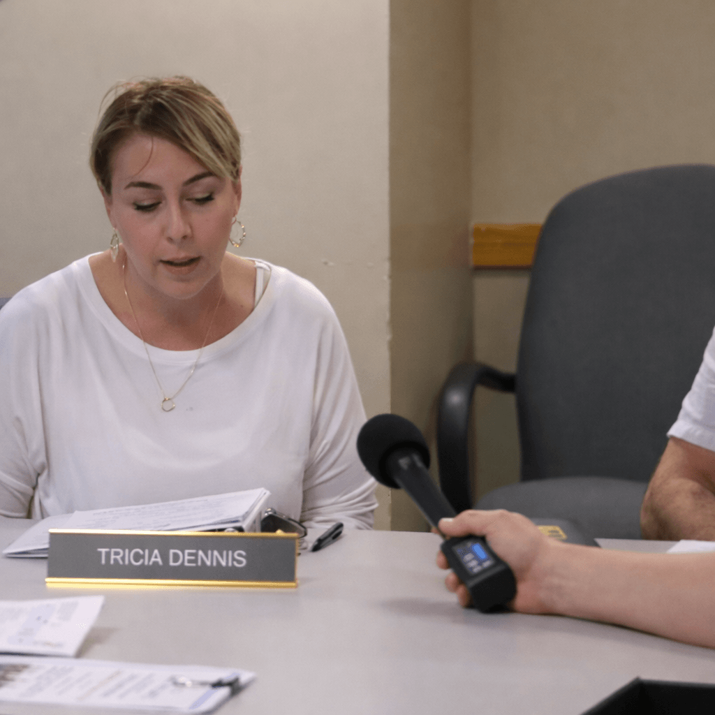 Woman with short hair reads papers at a table during an interview, with a microphone held by another person.