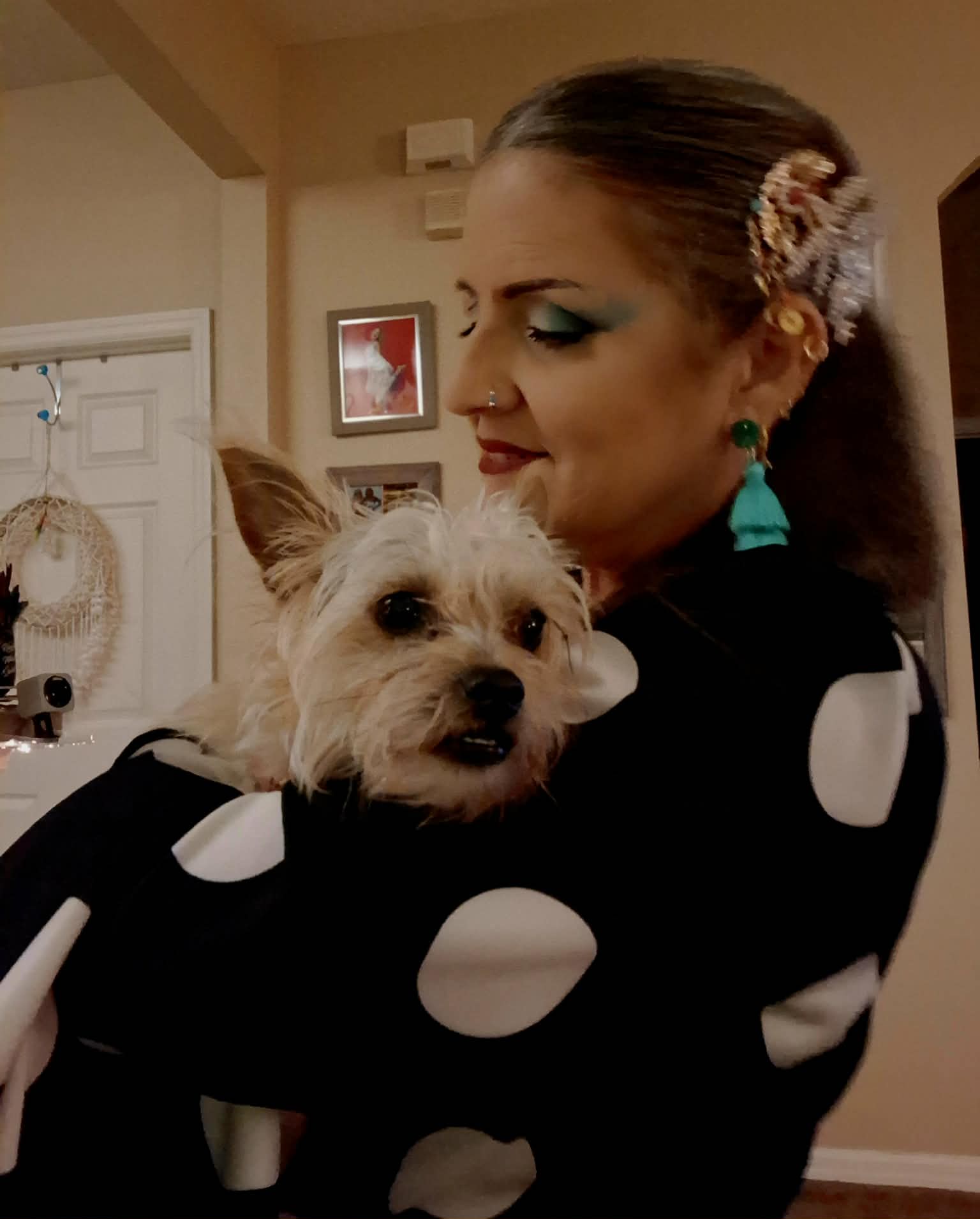 Woman holding a small dog with light fur, wearing a black dress with white polka dots, in a home interior.
