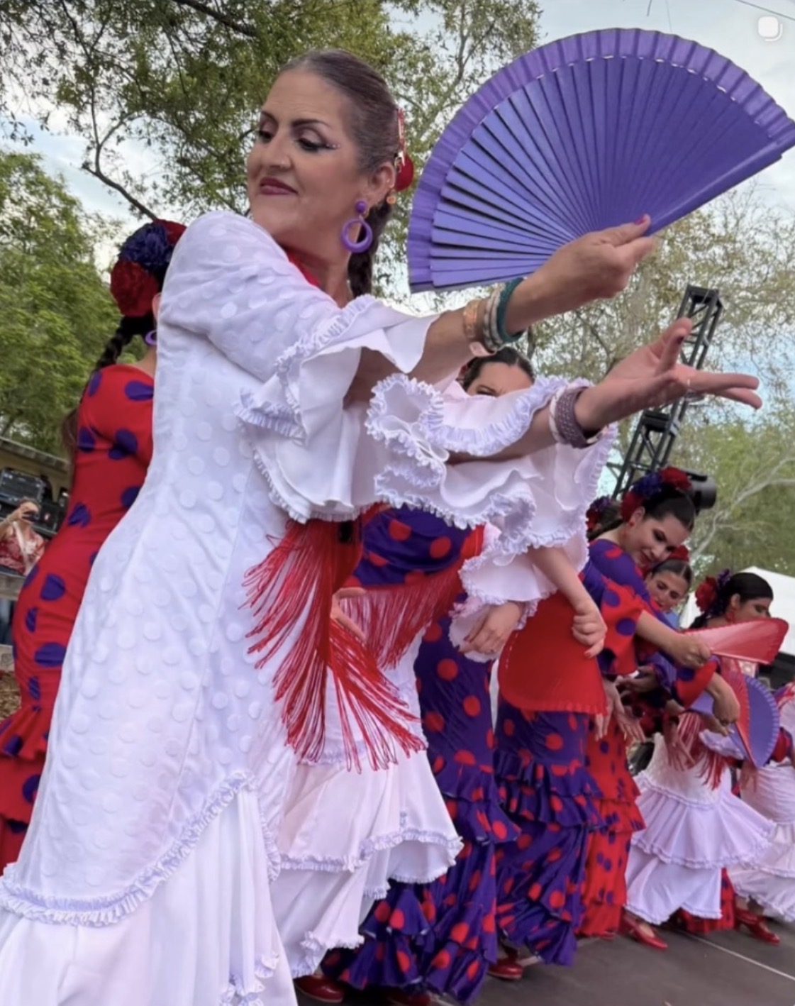 Group of women in traditional dresses dancing outdoors, one woman holding a purple fan, trees in background.