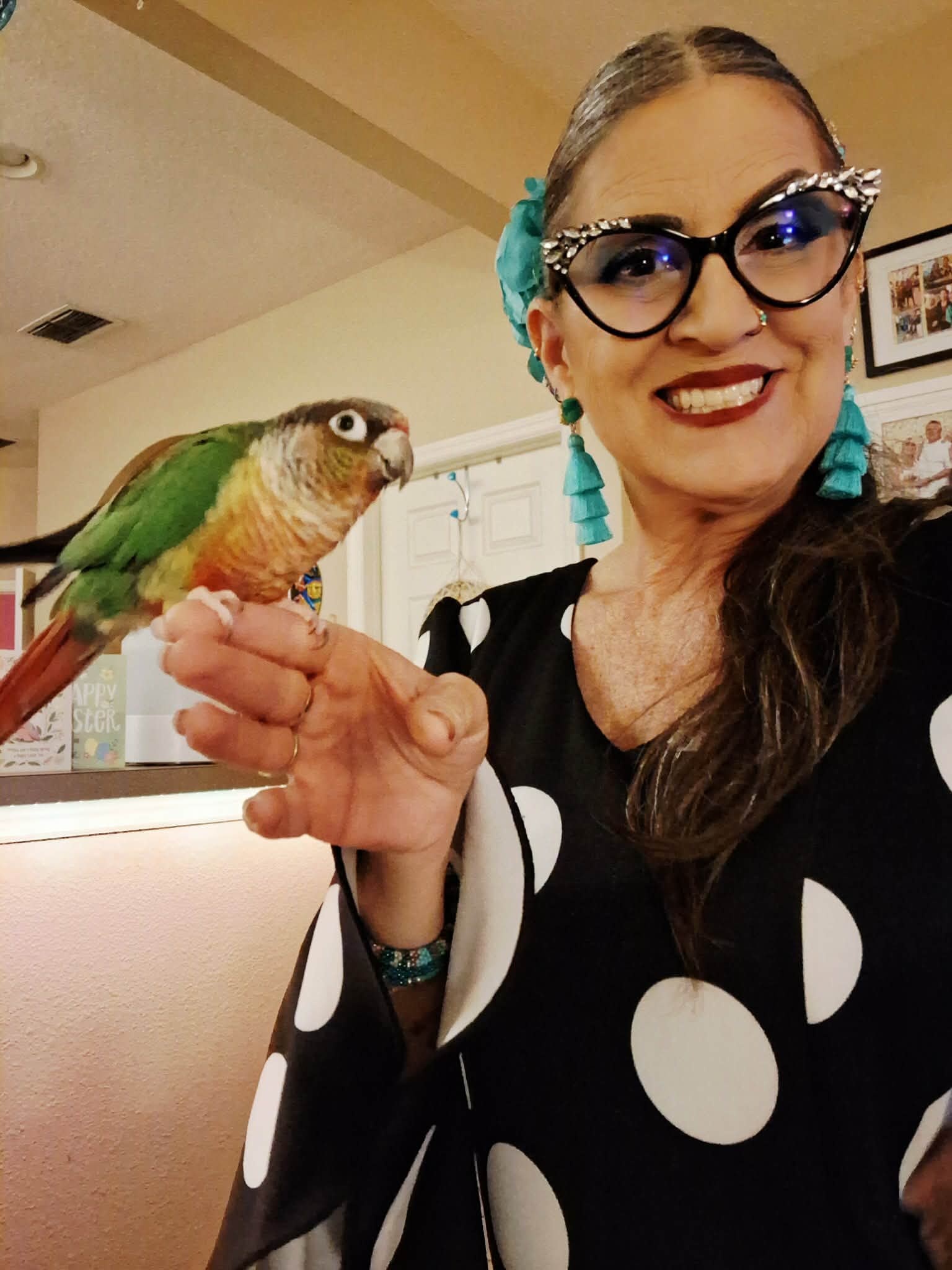 Woman with glasses and blue accessories holding a colorful parrot indoors.