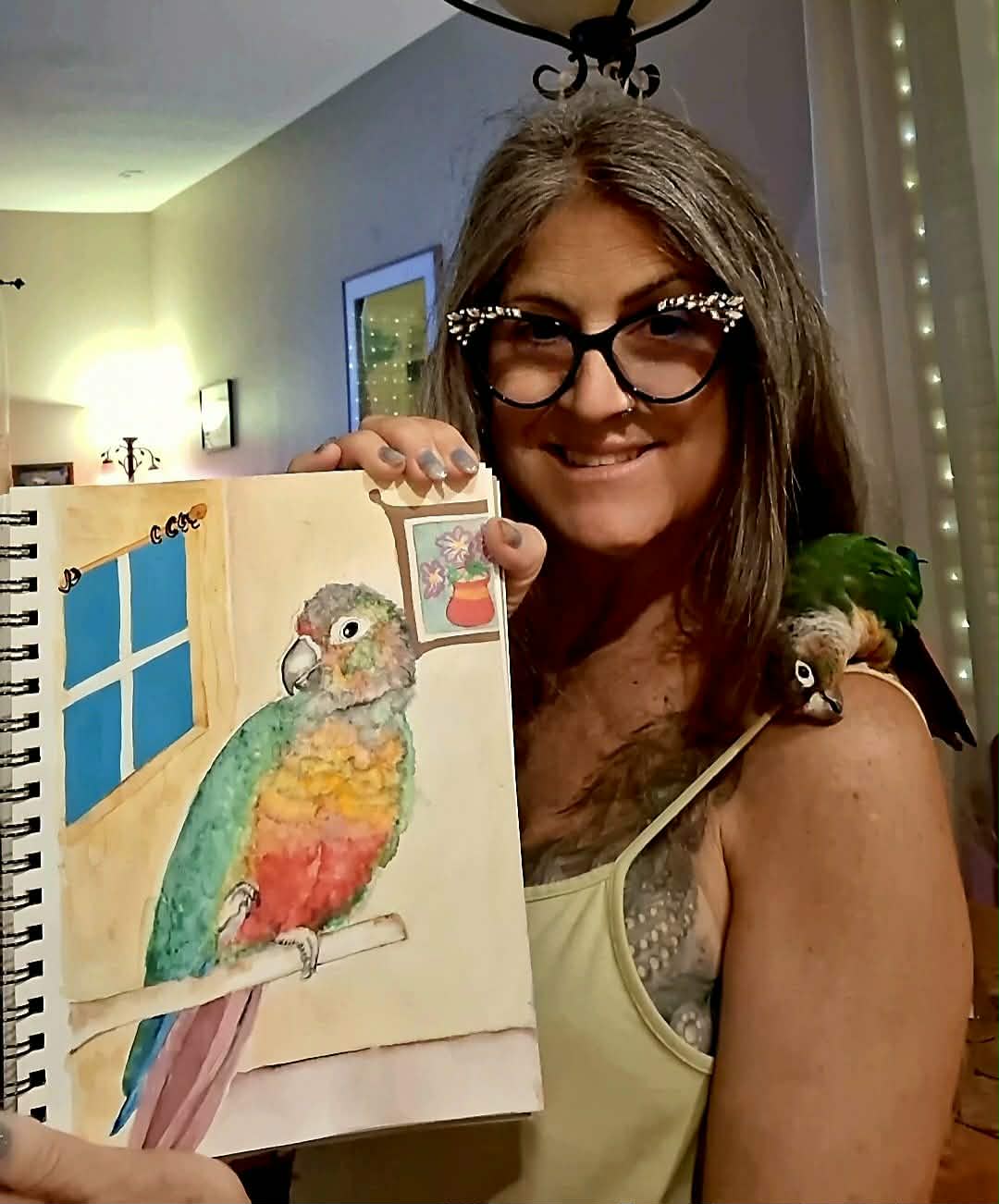 Woman with glasses smiling, holding a drawing of a colorful bird inside a room with a window and artwork on the wall.