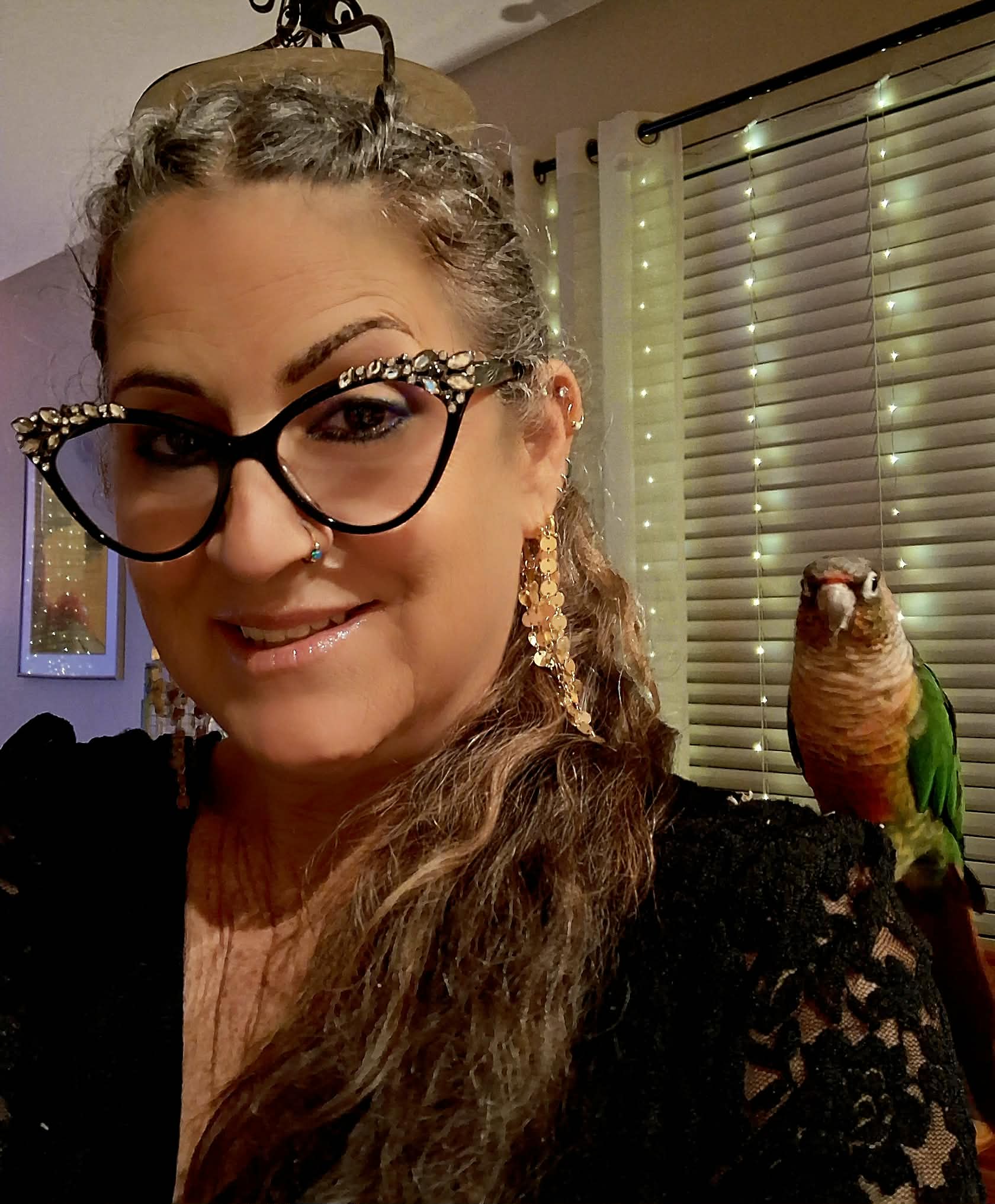 Woman with glasses and earrings, smiling, with a parrot on her shoulder, in a room with blinds and curtains.