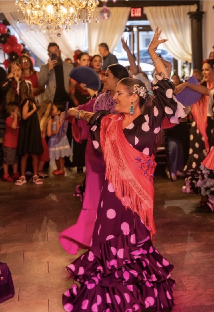 A woman in a pink and purple polka dot dress dances with arms raised at a festive event, surrounded by children and adults.
