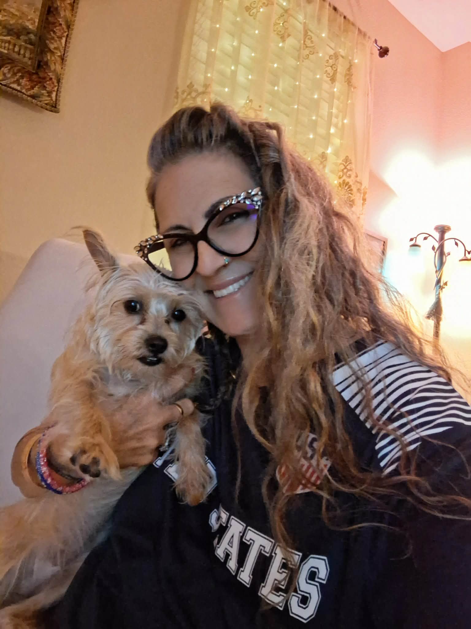 Woman with long curly hair and glasses holding a small dog indoors, smiling.