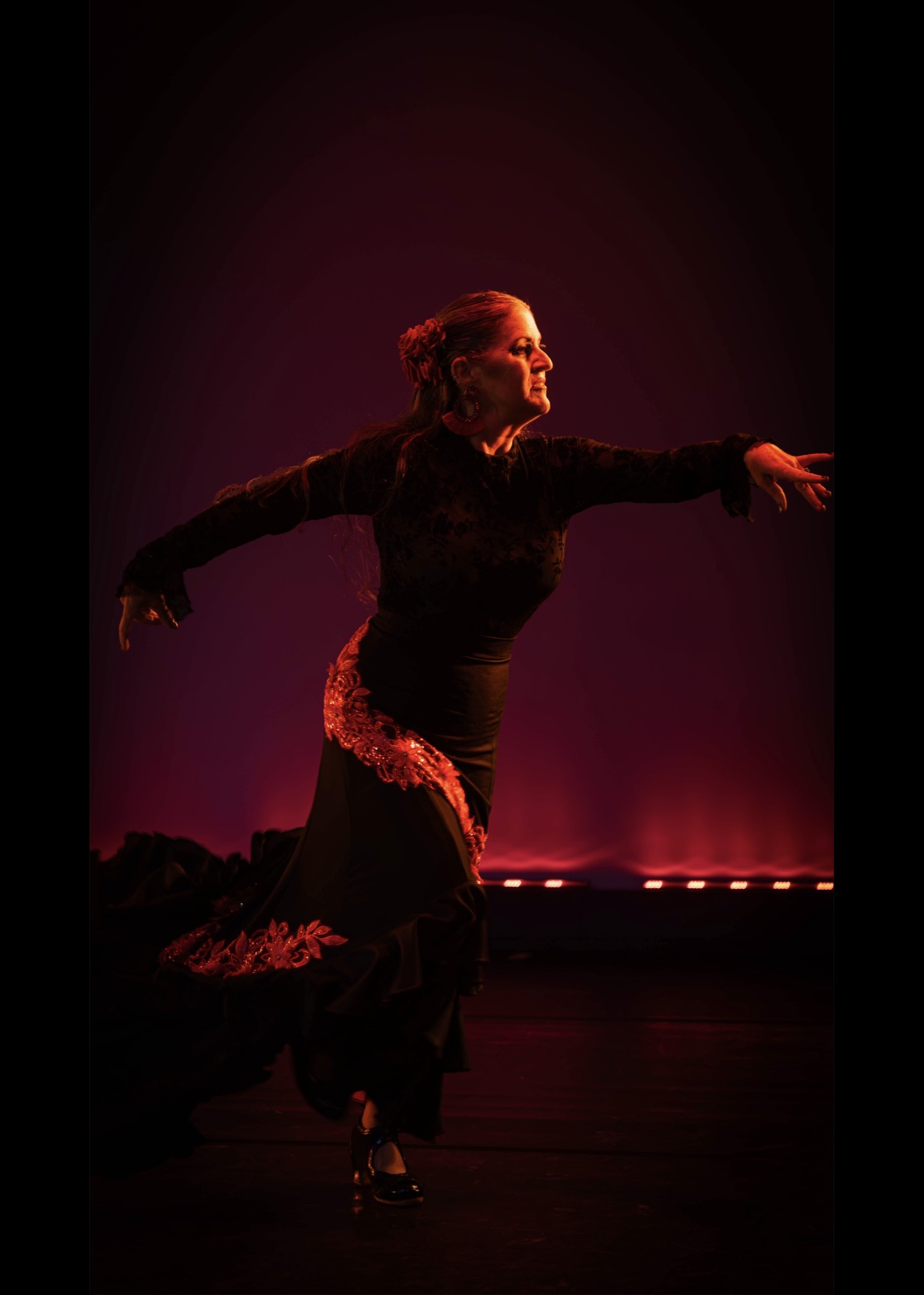 Performer dancing on stage with arms extended, illuminated by pink and purple lighting, wearing a black dress with red accents.