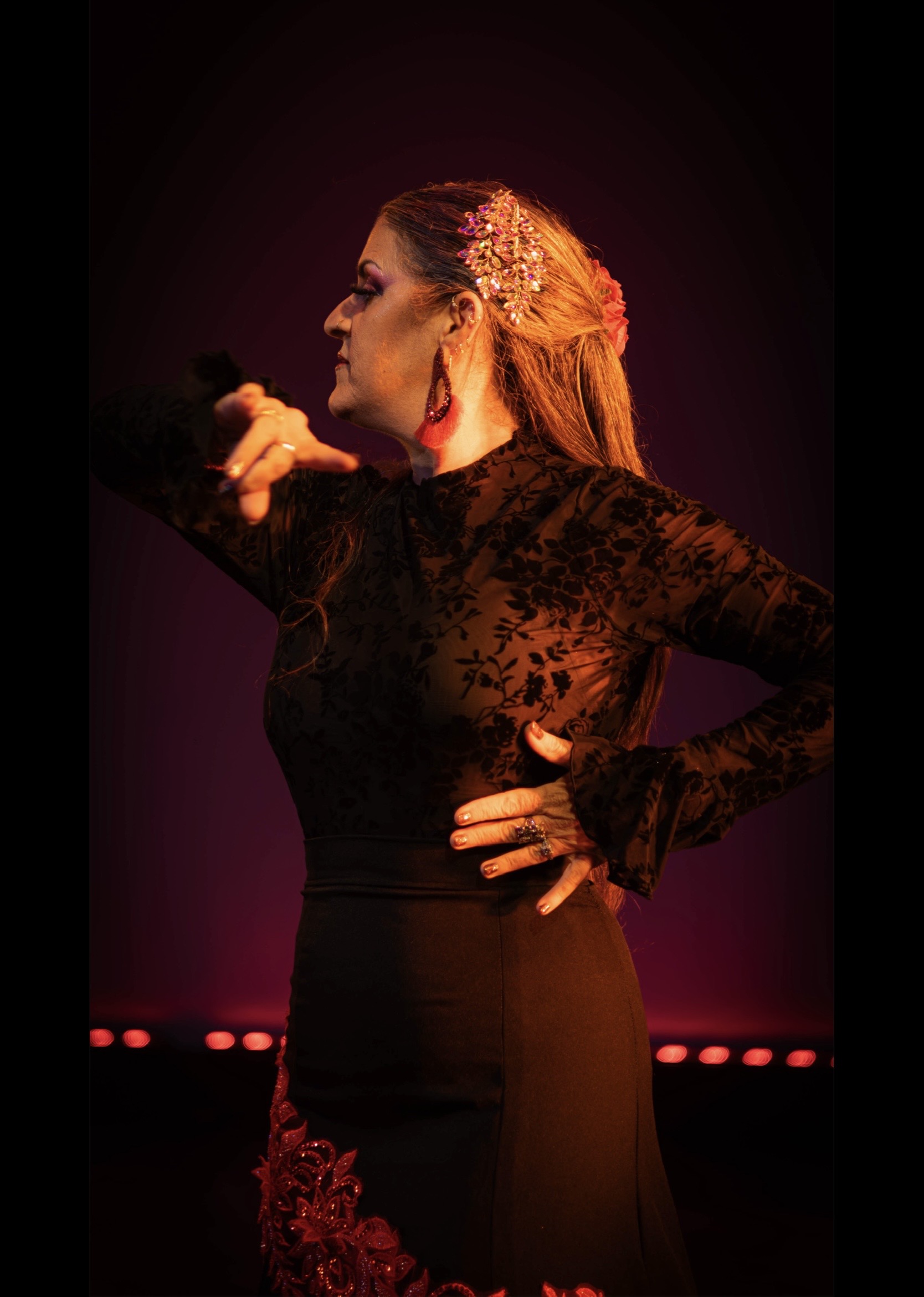 Side profile of a woman singing into a microphone, wearing a black lace dress, with stage lighting in the background.