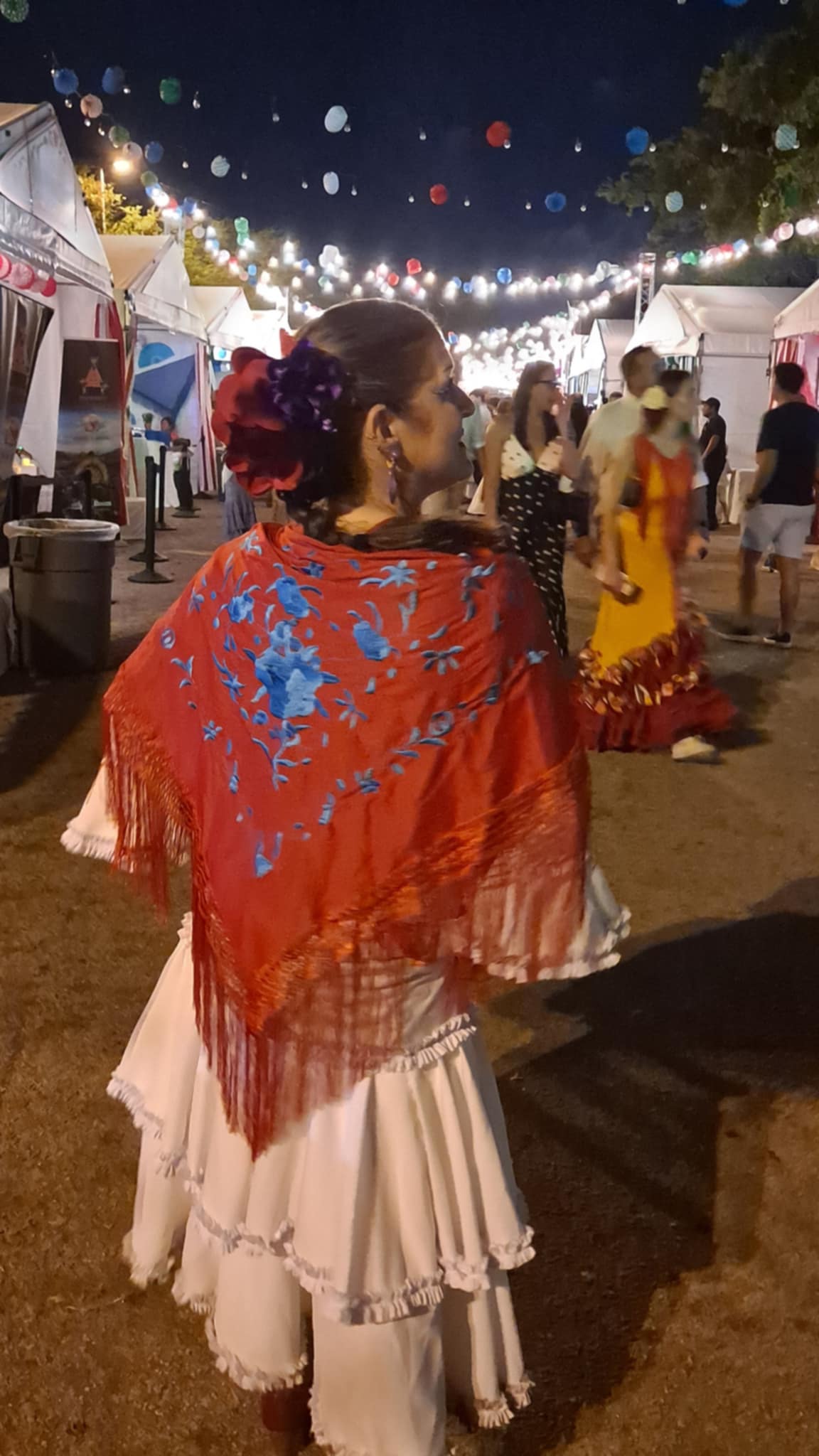 Woman in a red shawl and white dress walking at night festival with tents and string lights in background