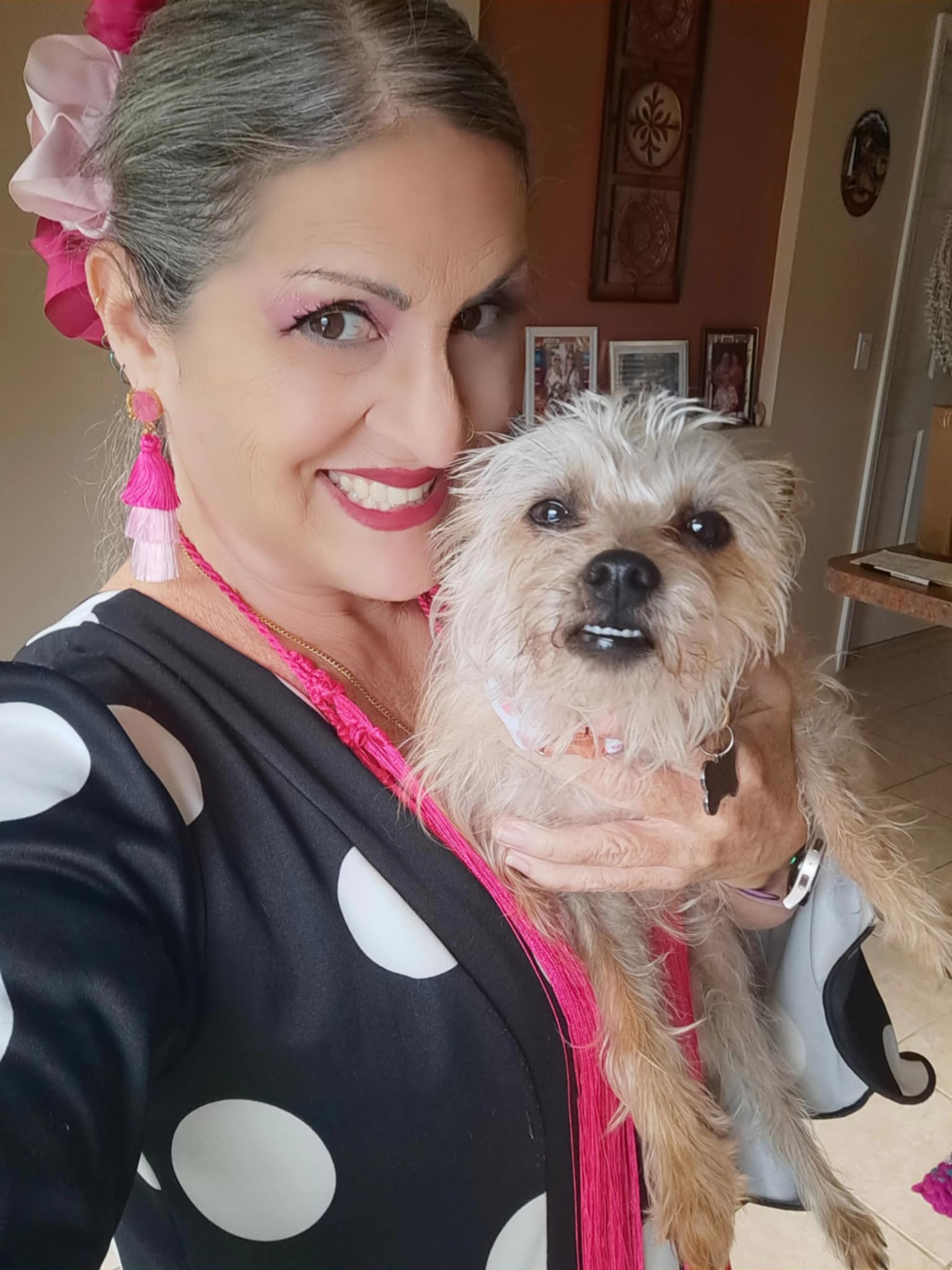 Woman smiling with a small dog indoors, background with framed pictures and wall decorations.
