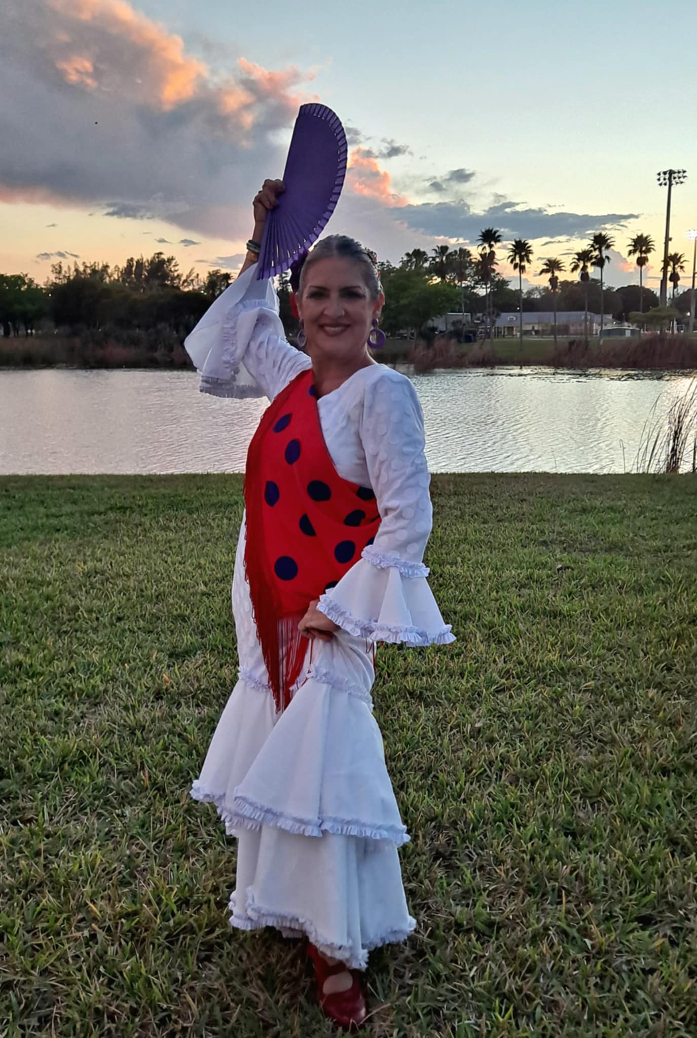 Woman in traditional dress holding a purple fan outdoors near water, with trees and sky in background.