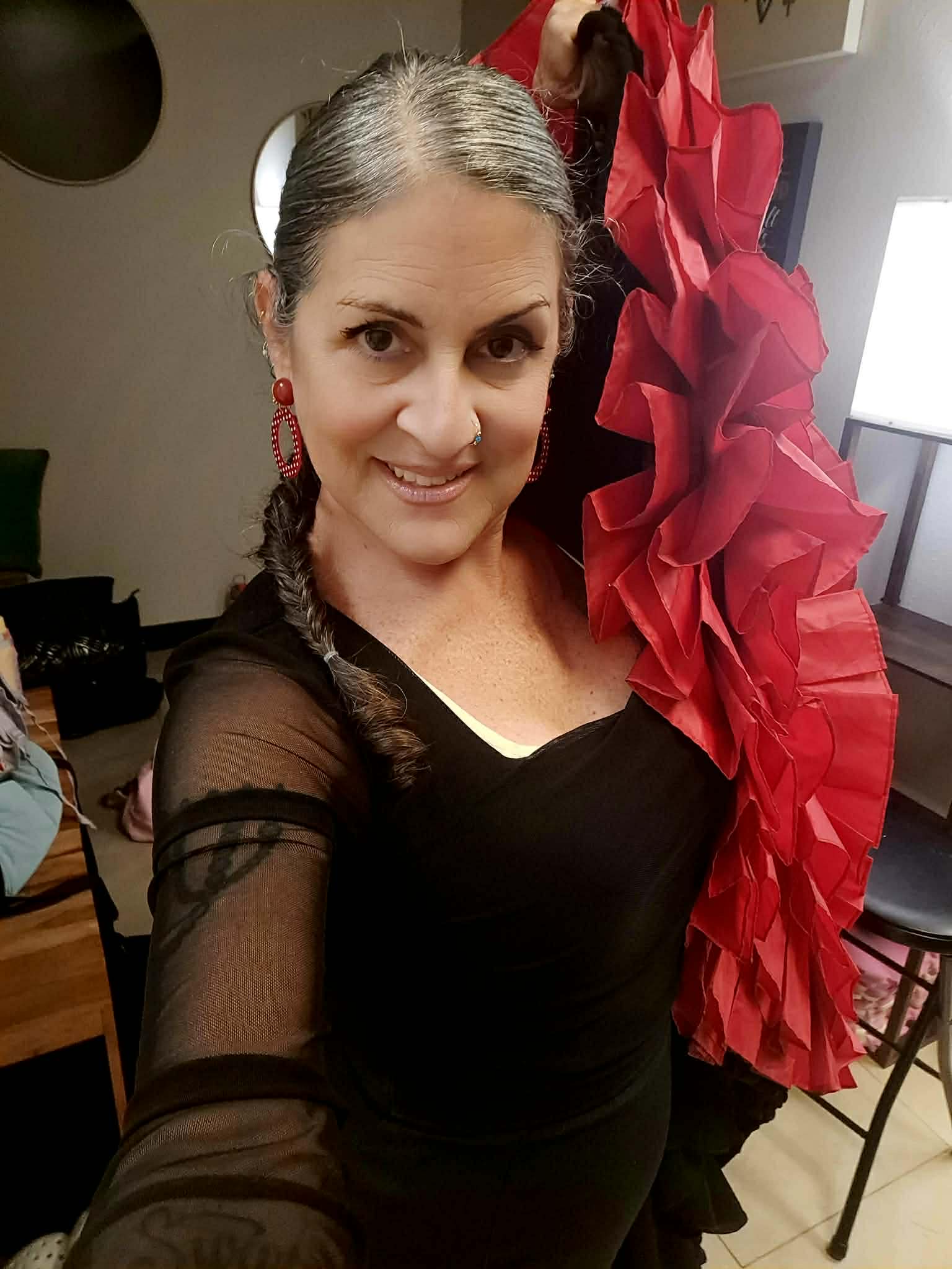 Woman with braided hair smiling, wearing black dress with sheer sleeves, holding large red flower decoration, indoors.