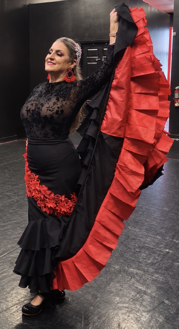 Woman in black dress holding a large red and black ruffled skirt, smiling with eyes closed, in a dance pose.