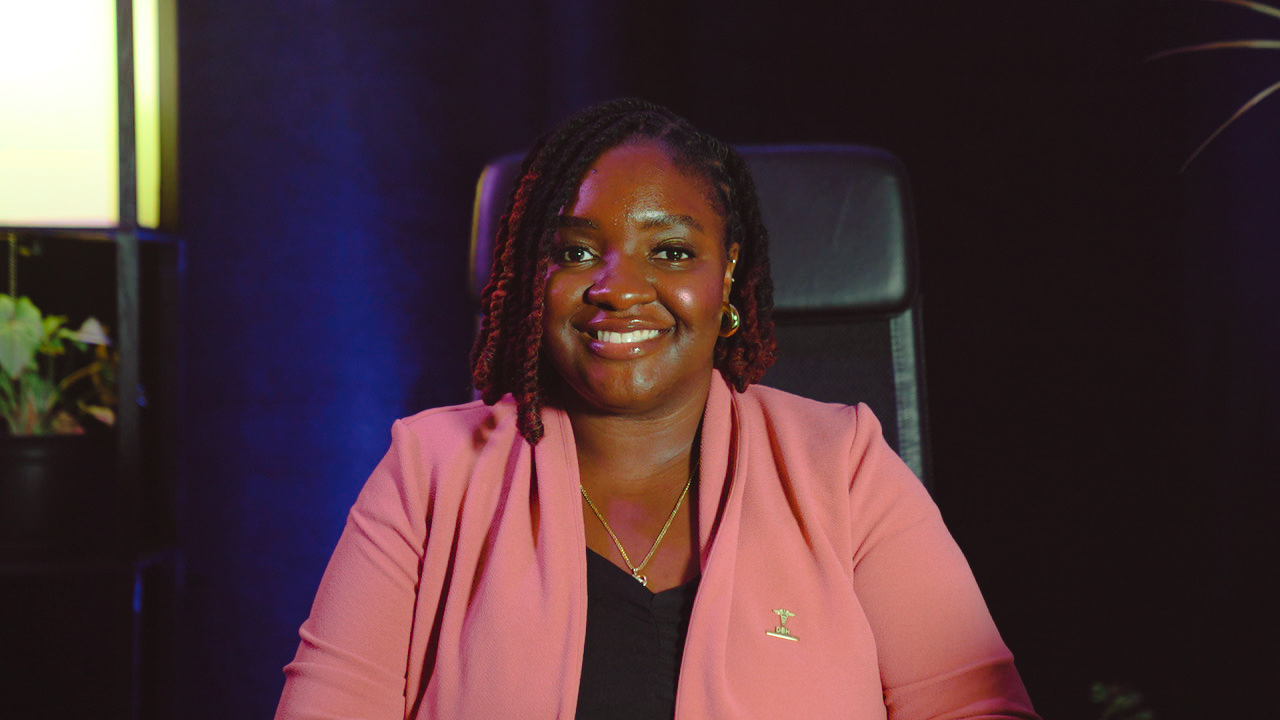 Woman with braided hair smiling, wearing a pink blazer, sitting in a dark room with a black chair.