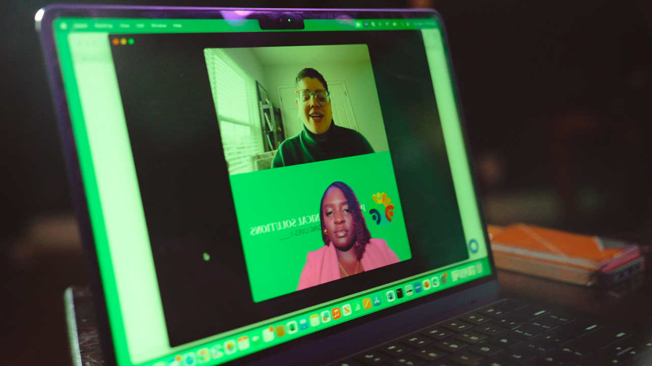 Laptop screen showing a video call with two people, one smiling and one with a neutral expression, in a bright room.