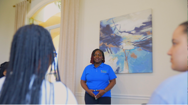 A woman in a blue shirt stands in front of a group, with a painting on the wall behind her.