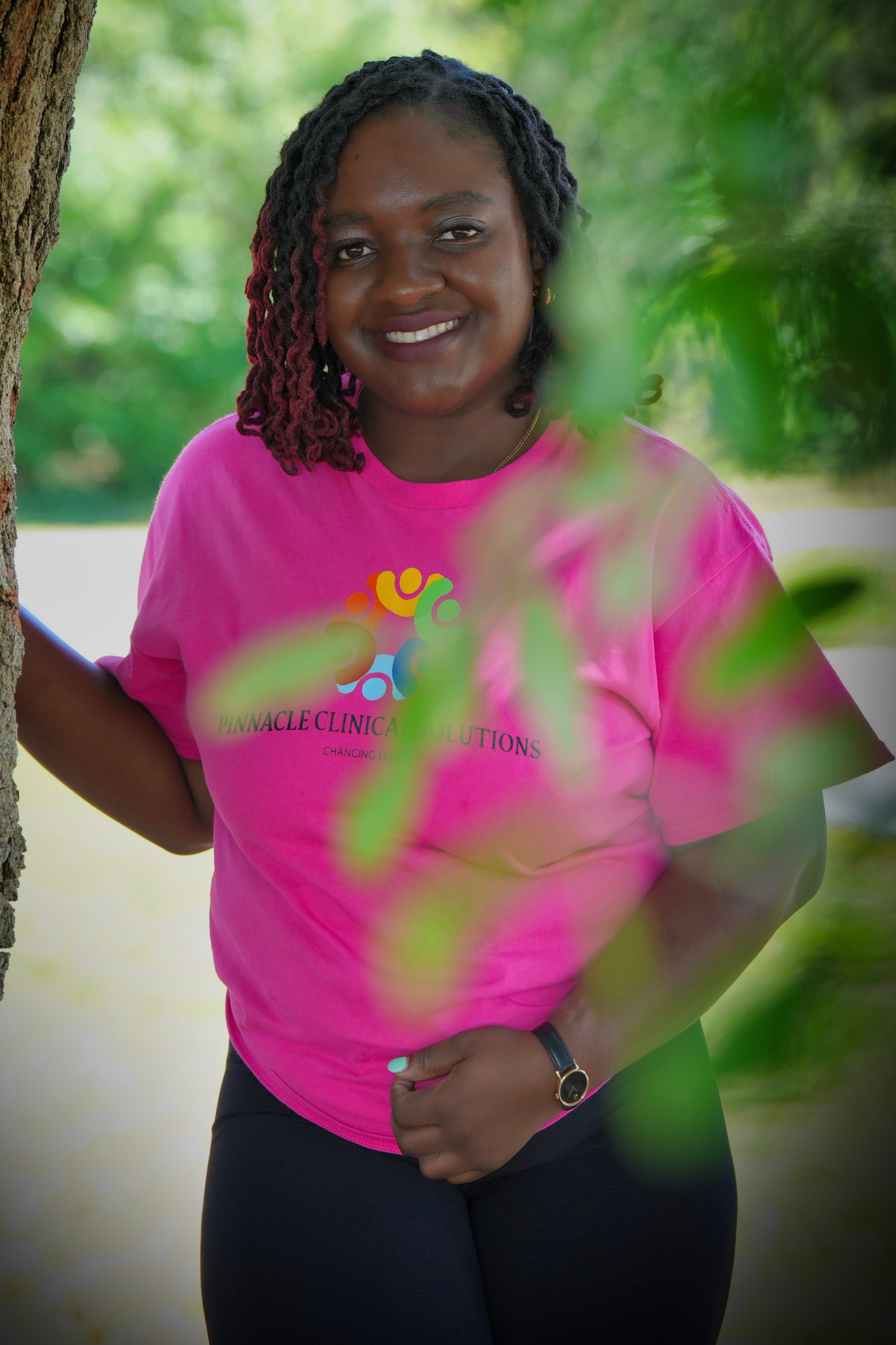 Woman with braided hair smiling outdoors, wearing a pink T-shirt and black pants, surrounded by greenery.