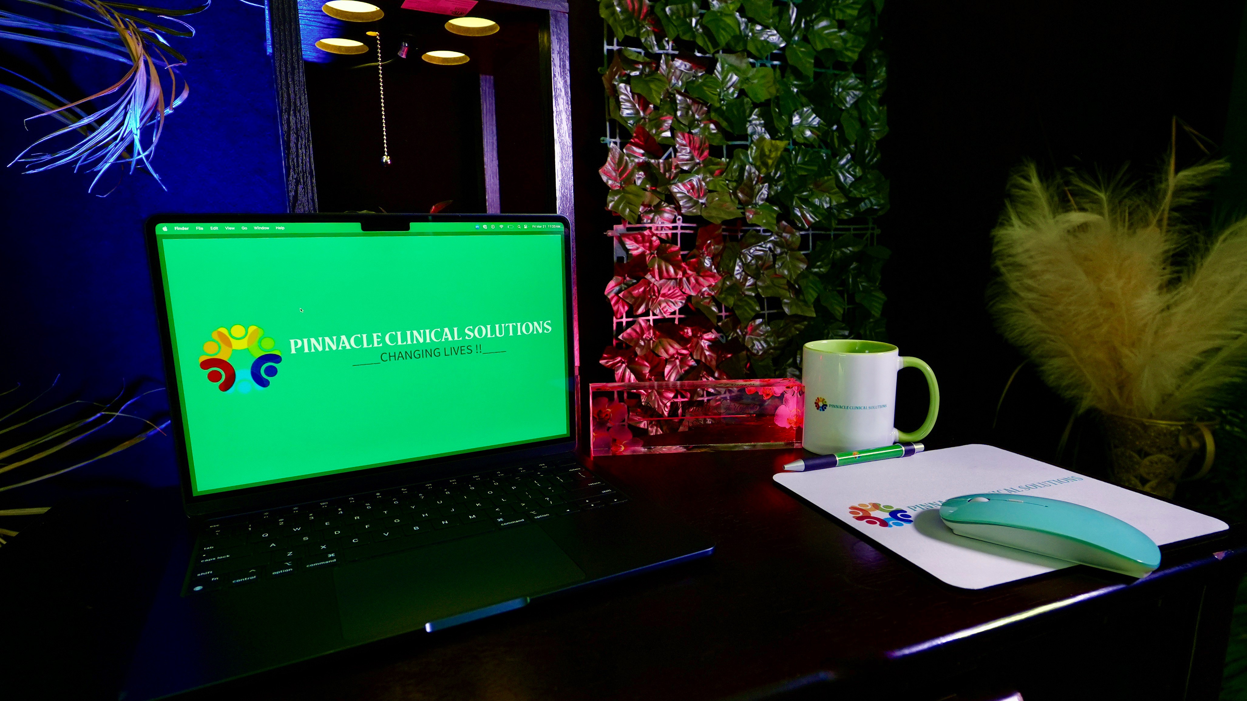Laptop, mug, notebook, and pen on a desk with plants and flowers in the background.