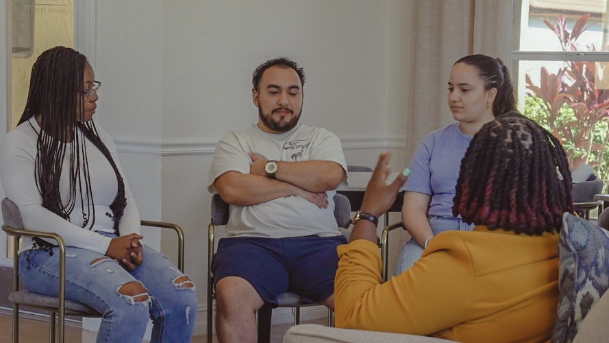 Four people sit in a circle having a discussion in a room with a window and plants outside.
