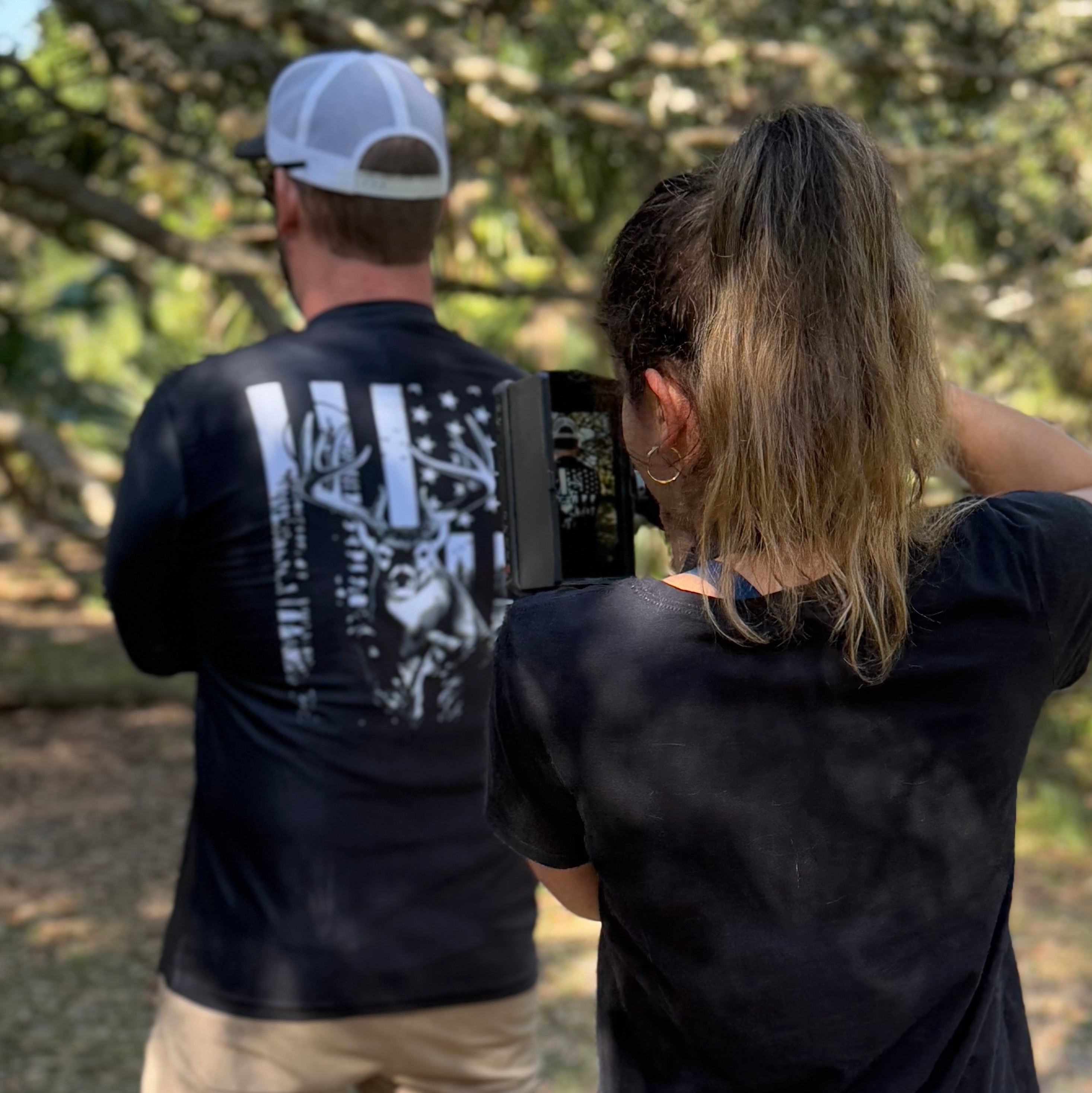 Two people outdoors, one taking a photo or video with a smartphone, trees in the background, sunlight filtering through leaves.