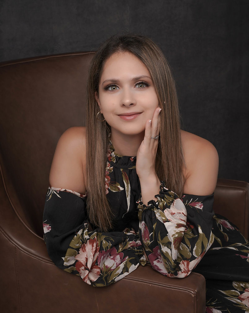 Young woman with long brown hair, smiling, resting her face on her hand, wearing a floral off-shoulder top, sitting on a brown chair.