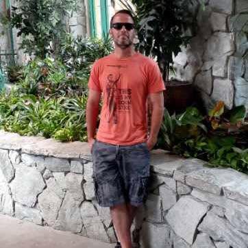 Man wearing sunglasses, red T-shirt, and cargo shorts standing outdoors near a stone wall with greenery.