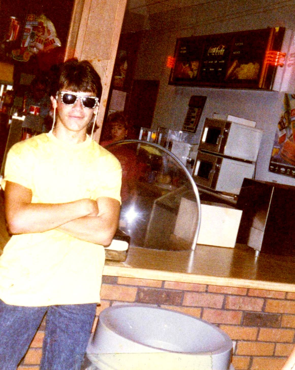 Person with dark hair, sunglasses, yellow shirt, arms crossed, standing in a room with kitchen appliances and brick wall.