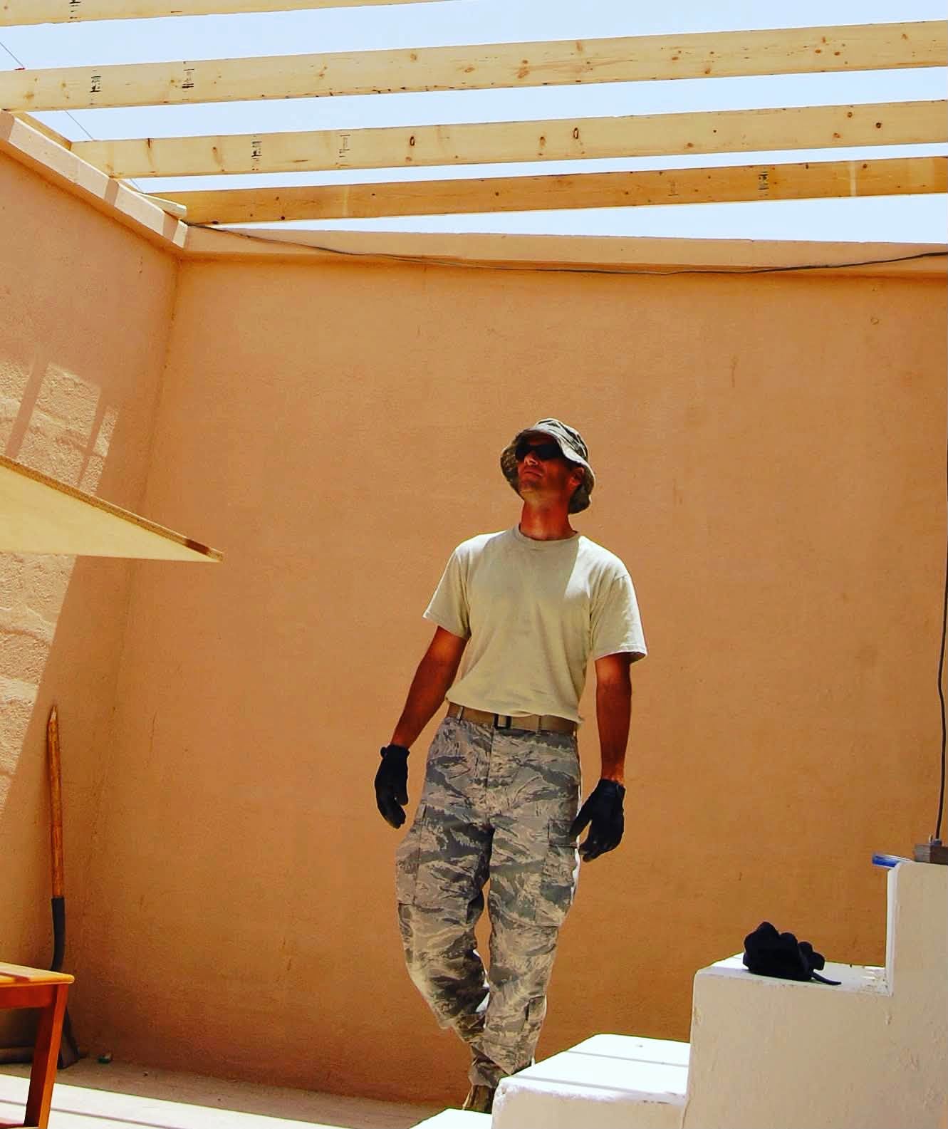 Person standing inside a partially constructed building with a wooden roof frame, wearing a helmet, mask, gloves, and camouflage pants.