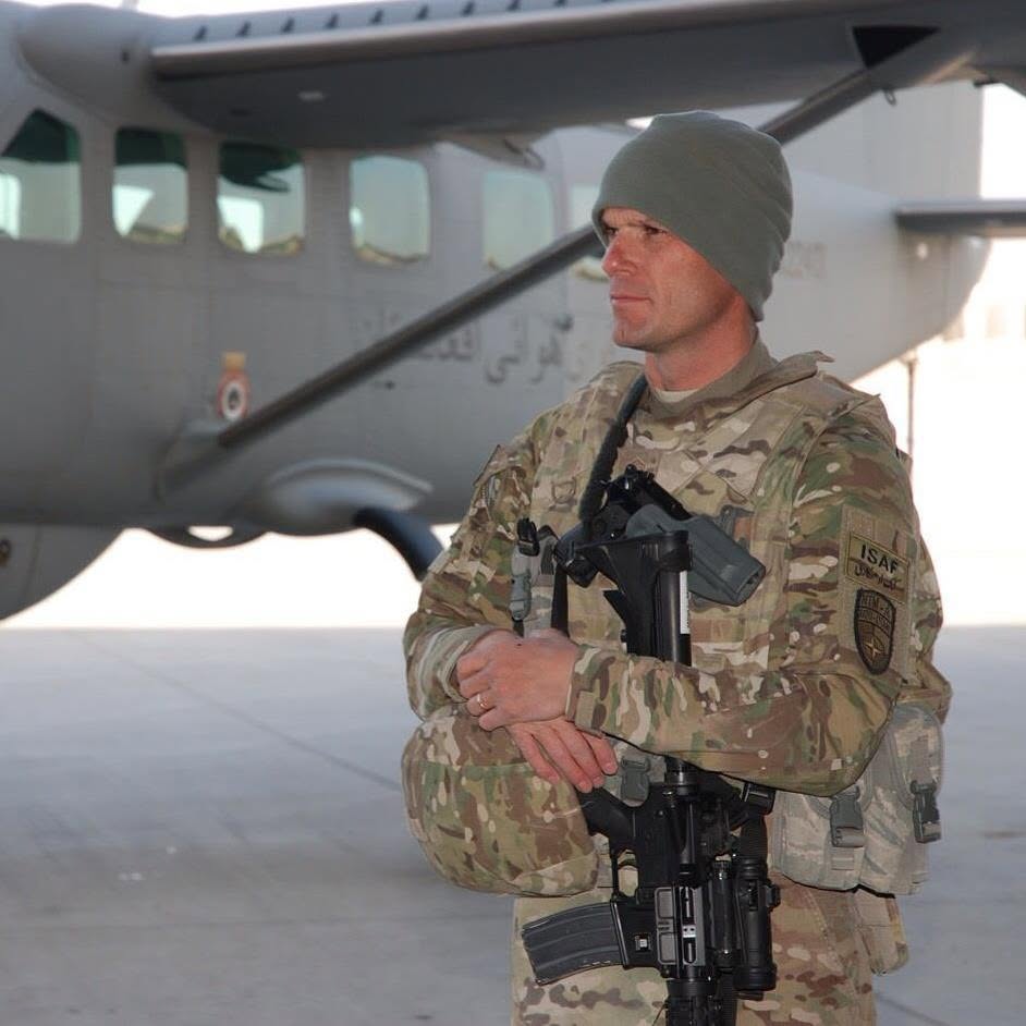 Soldier in camouflage uniform and gray beanie holding a rifle, standing near an aircraft on tarmac.