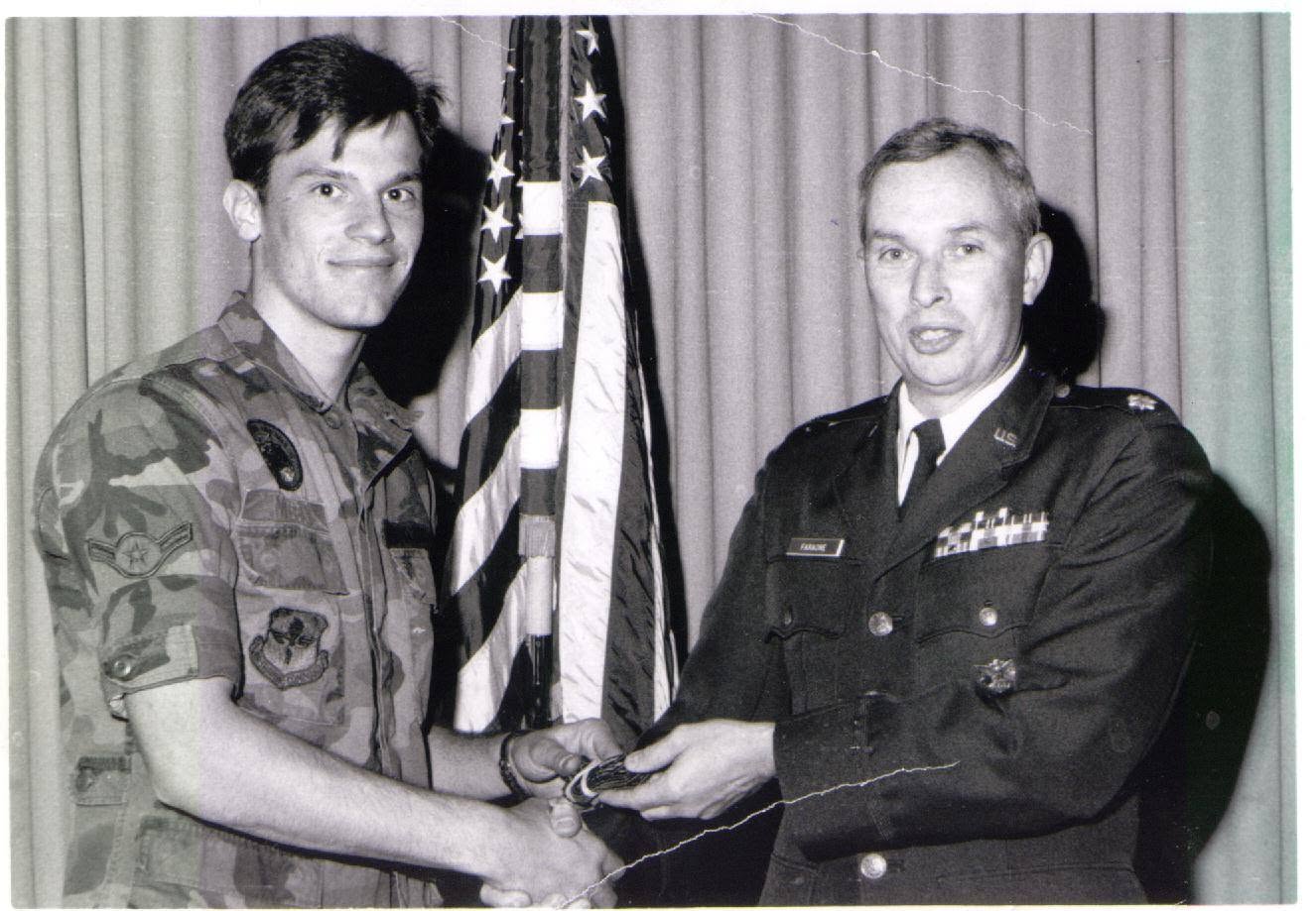 Two men in military uniforms shaking hands, with an American flag and curtain in the background.