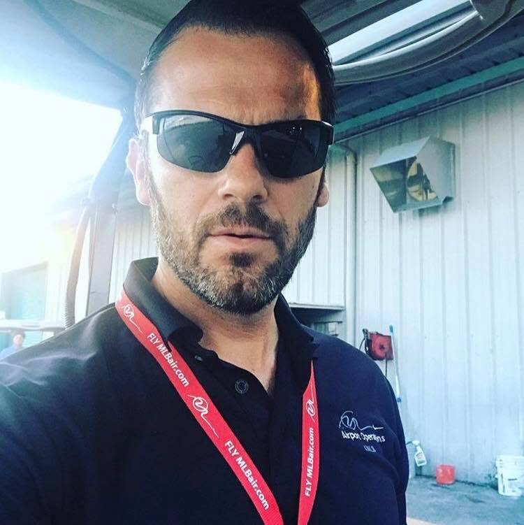 Man with dark hair, beard, sunglasses, wearing a black polo shirt with logos, outdoors near a building.