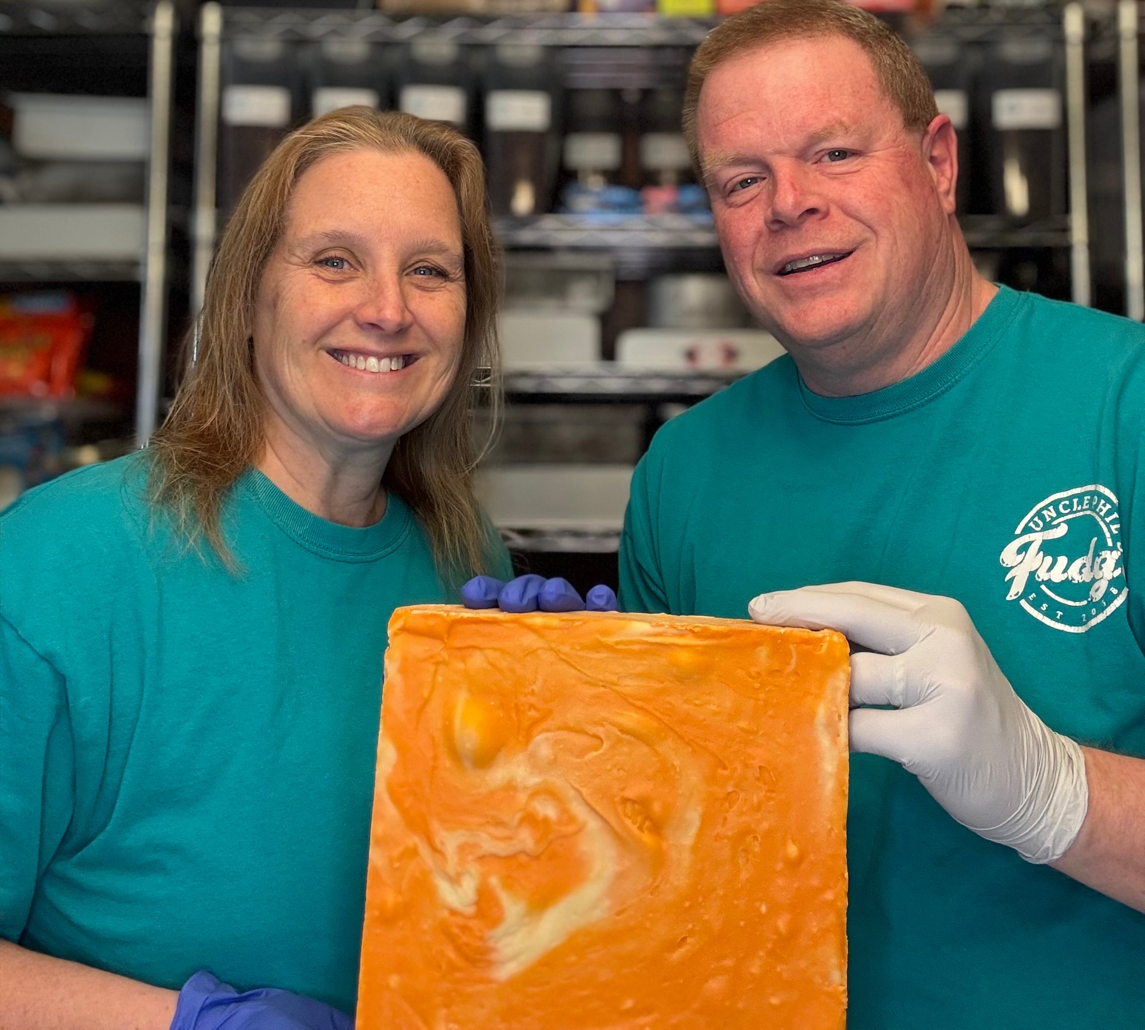 Two people in teal shirts holding a large orange soap bar, smiling, in a workshop setting.