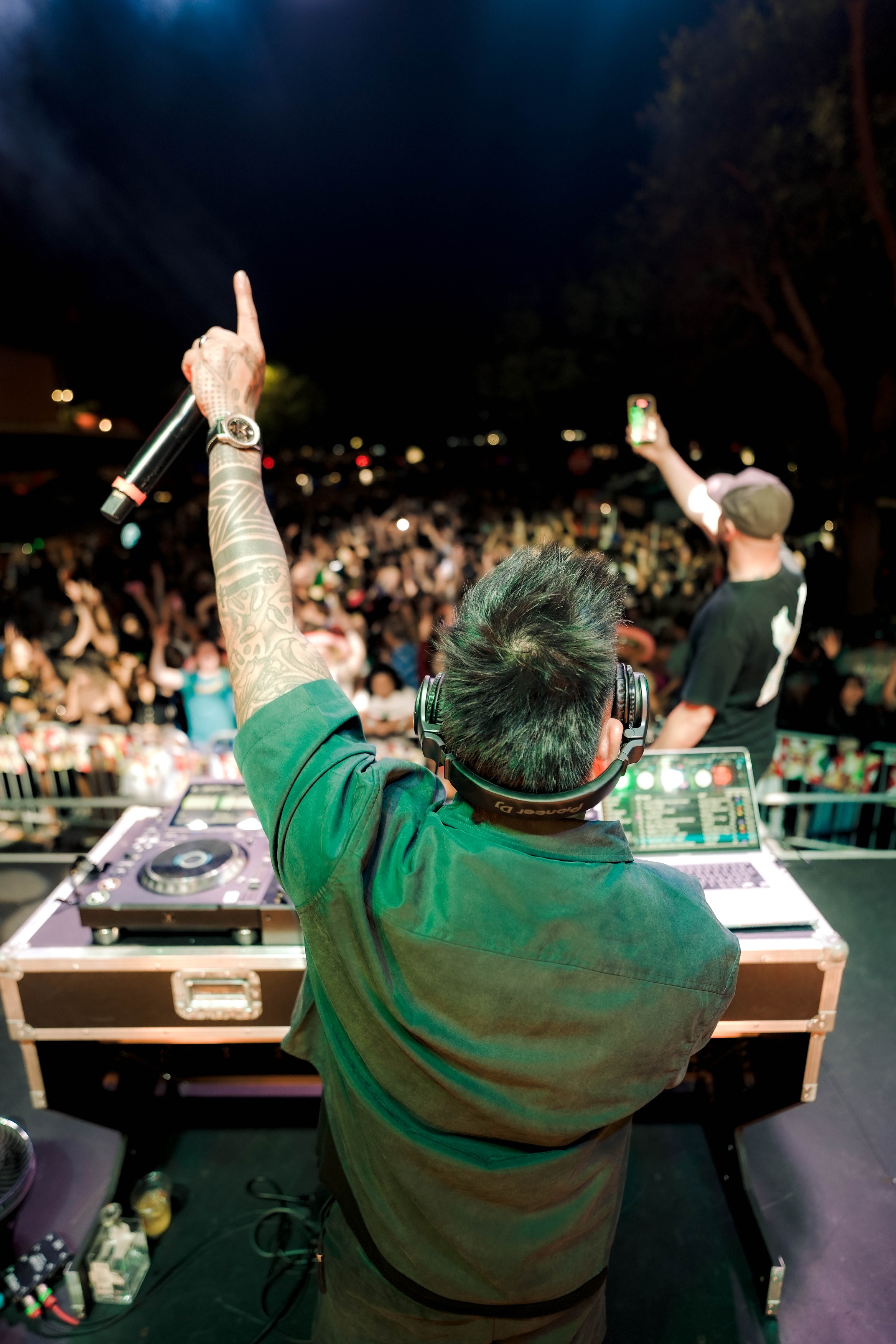 DJ with green hair and green shirt performs at outdoor concert, crowd in background, night sky visible.