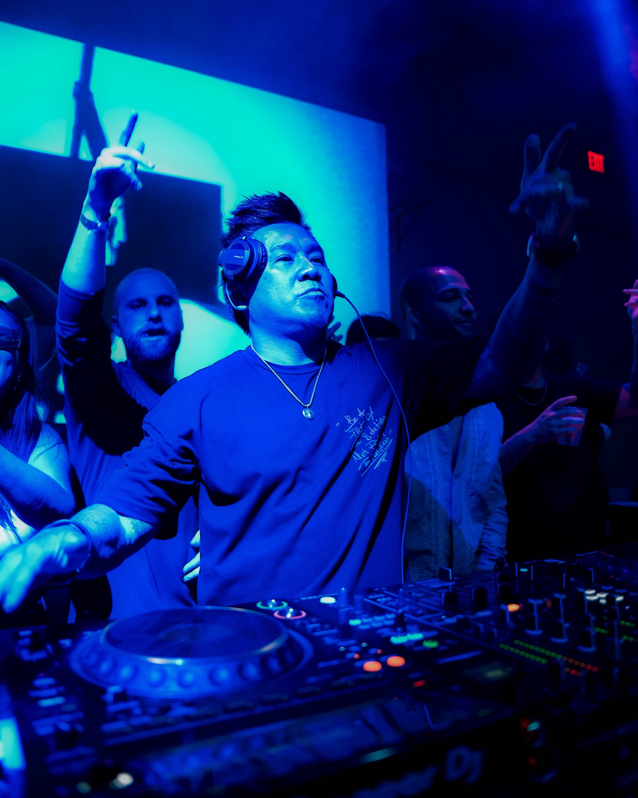 DJ with headphones at a club, raising hands, surrounded by people, blue lighting, DJ equipment visible