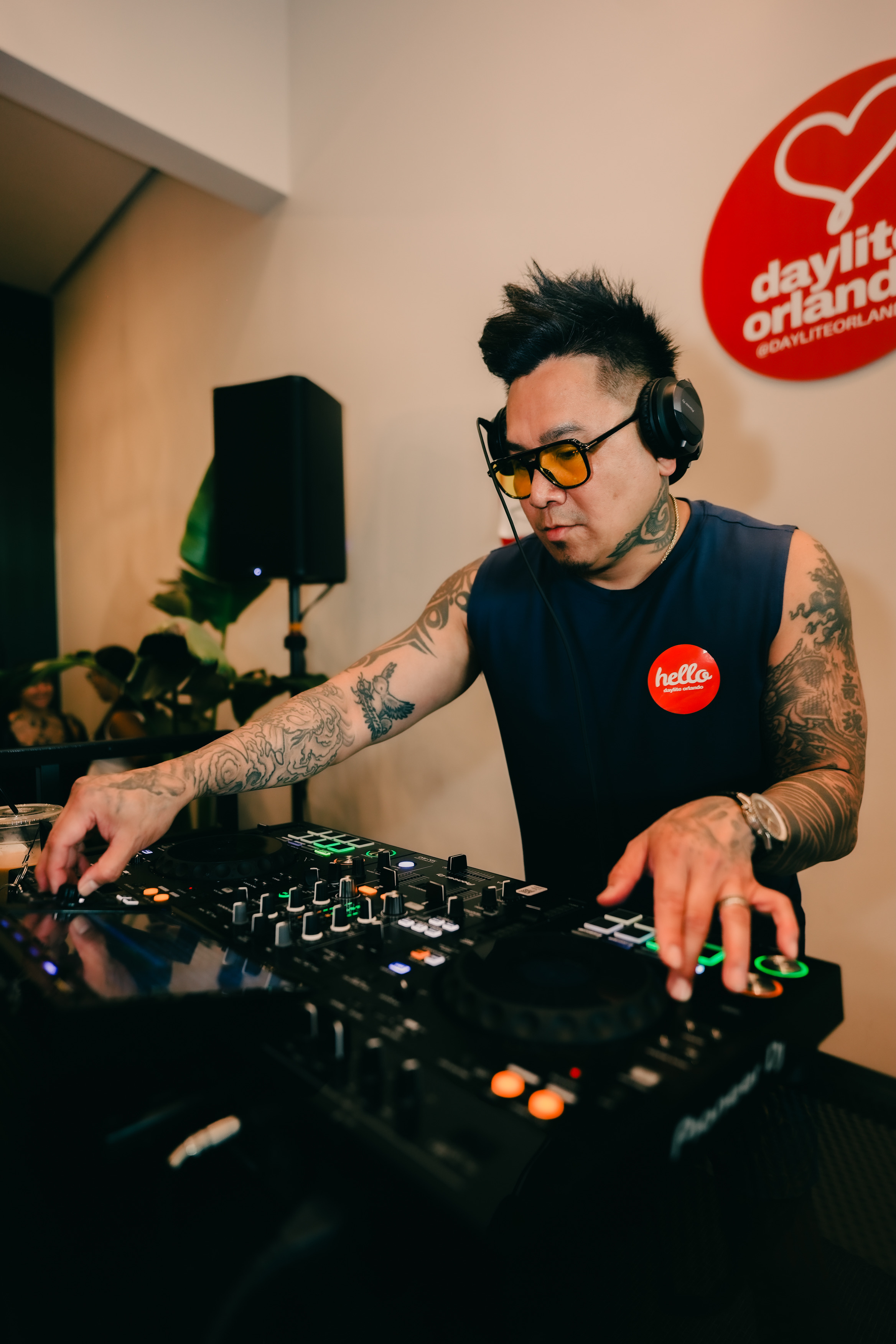 DJ wearing headphones and sunglasses, operating a mixer in a studio with a red logo on the wall.