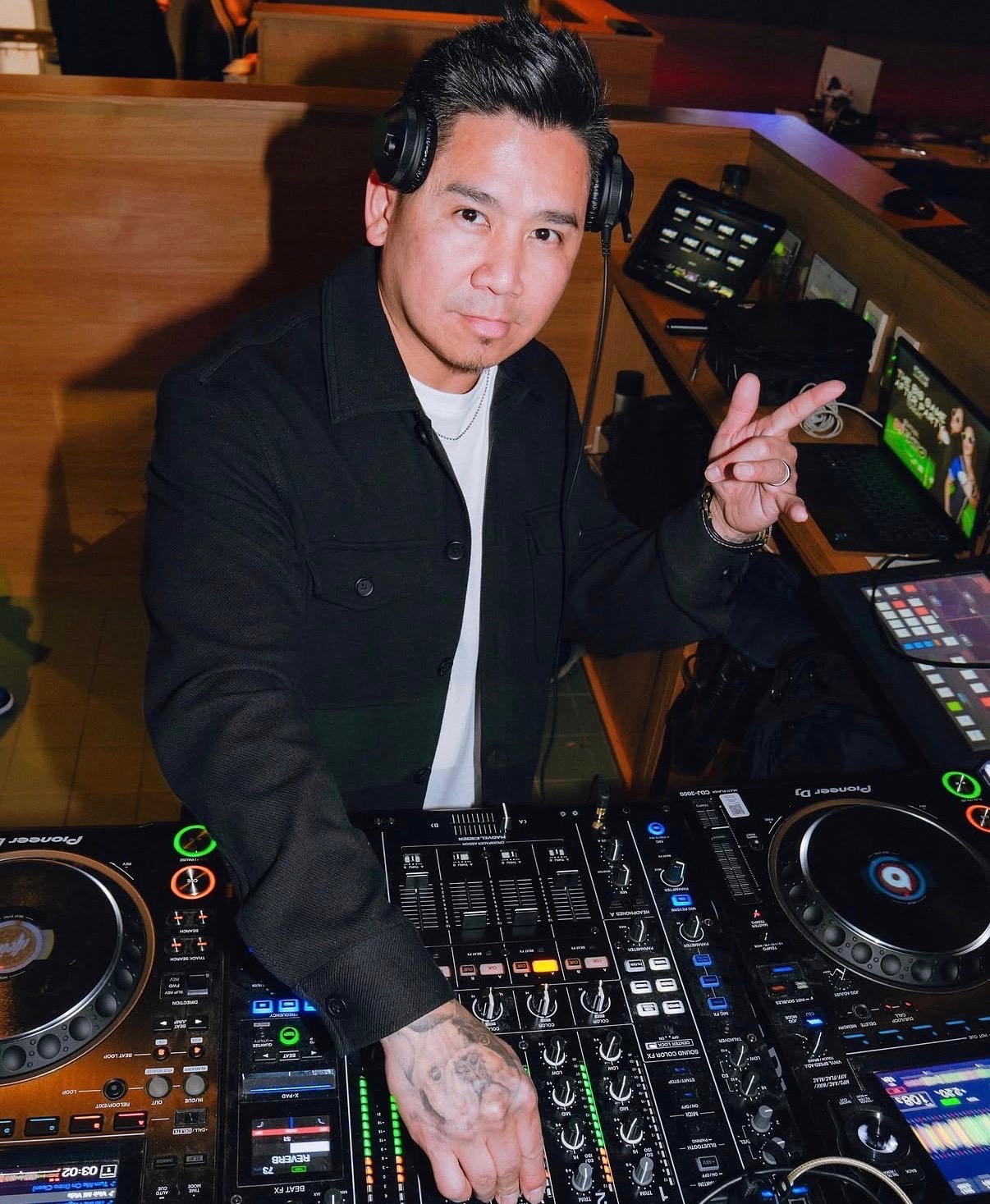Person with headphones making a peace sign, standing behind DJ equipment in a studio or club setting.