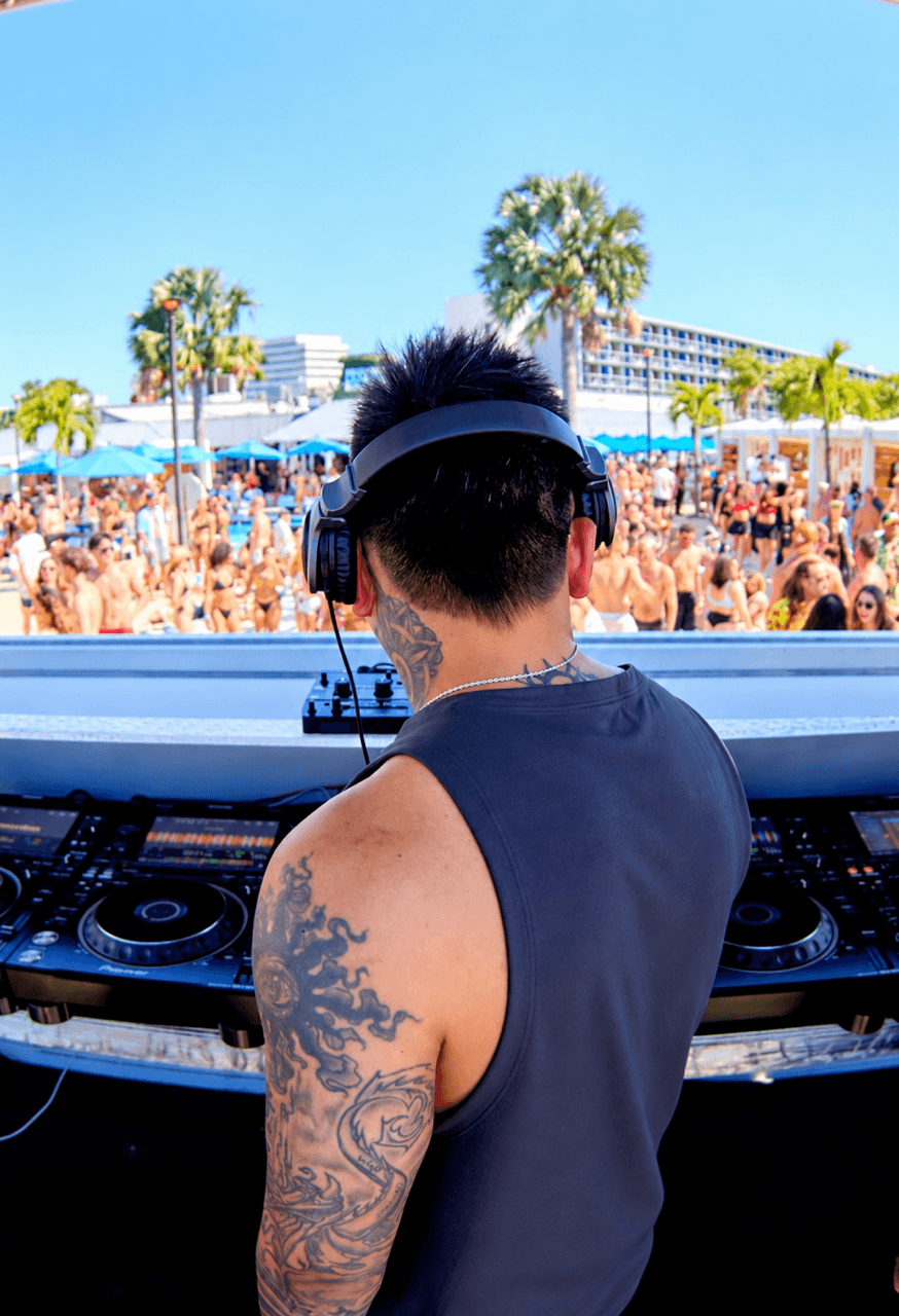 DJ wearing headphones and sleeveless shirt at outdoor event with crowd and palm trees in background