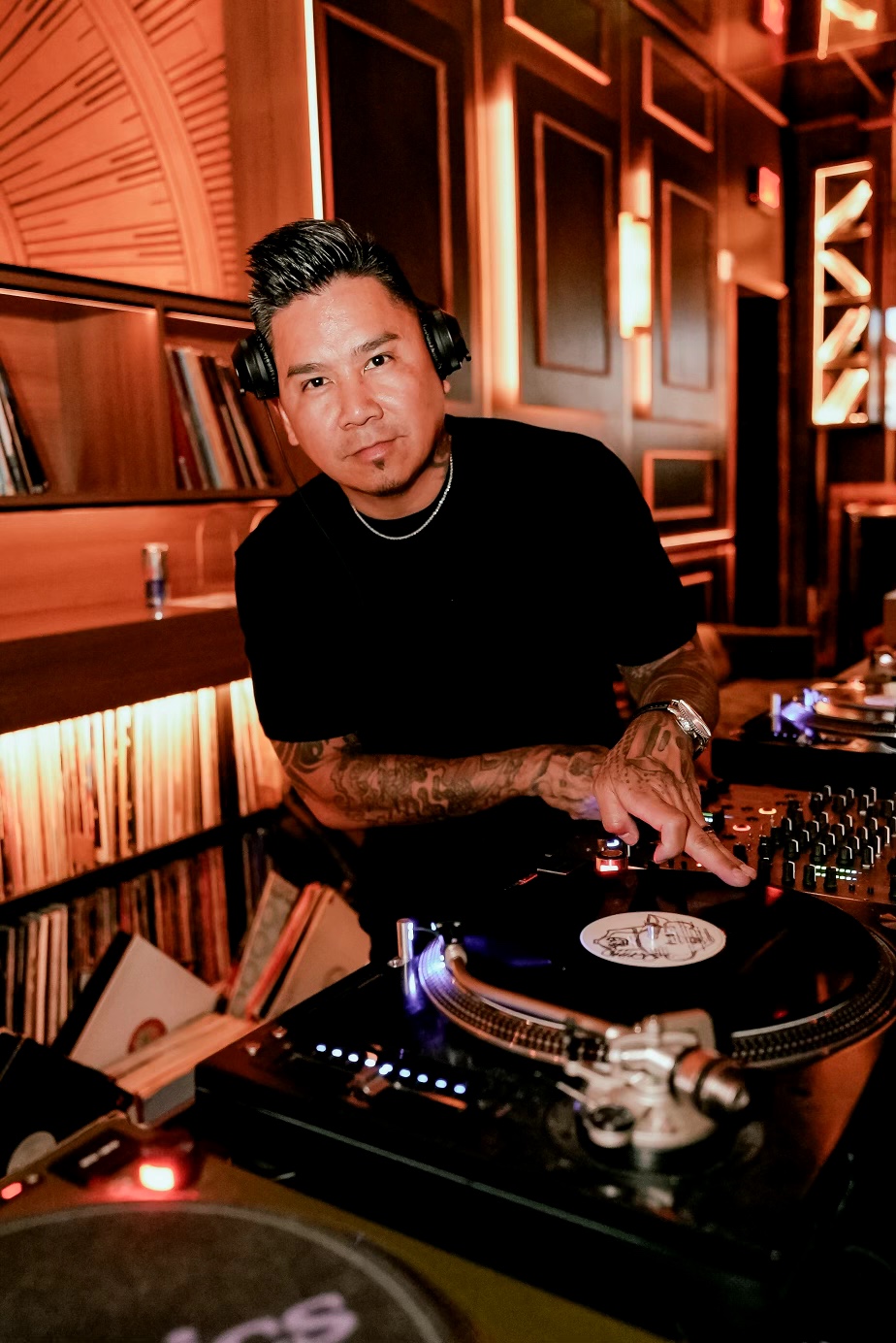 DJ with headphones operating turntable in a dimly lit, stylish room with wooden paneling and warm lighting.