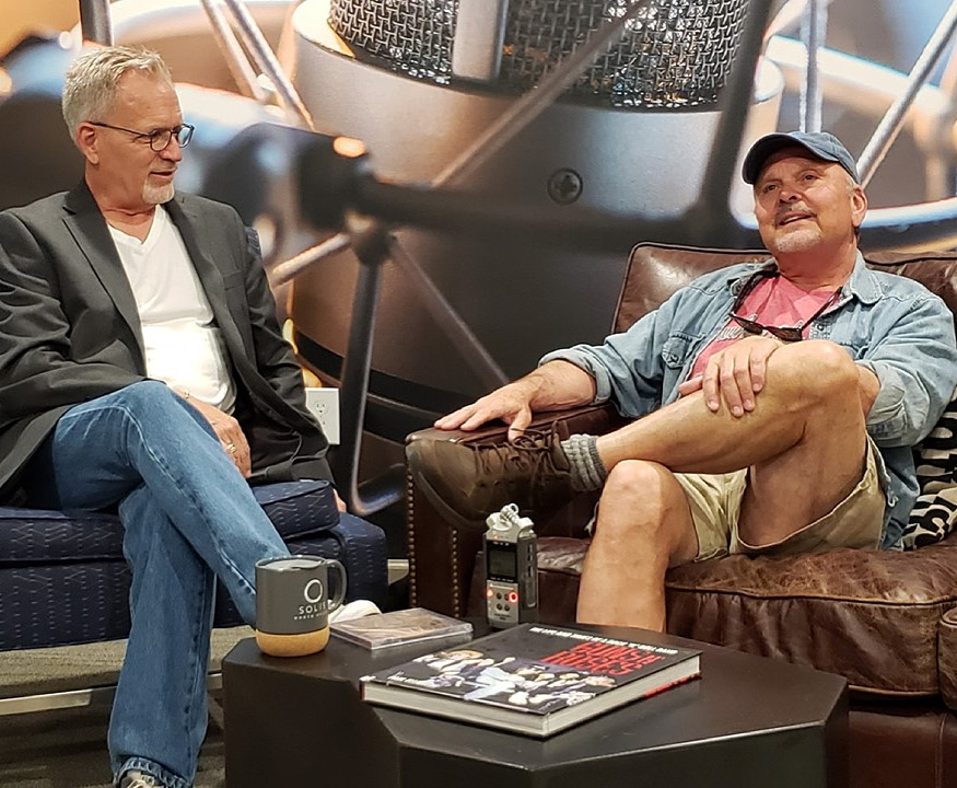Two men sit on couches in a studio setting with microphones and recording equipment visible, engaged in conversation.