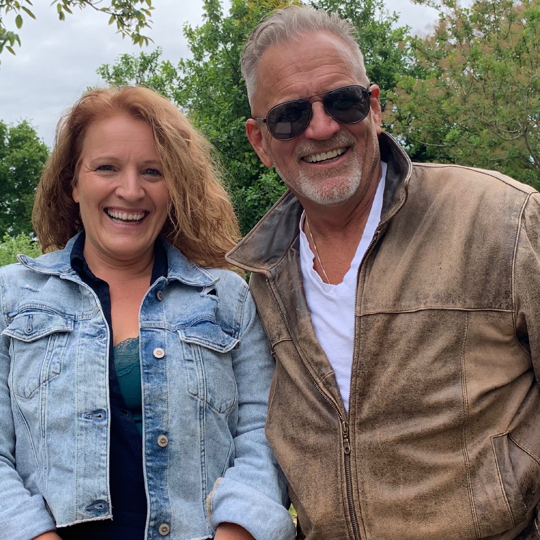 Two smiling people outdoors, woman with curly hair in a denim jacket, man with sunglasses and leather jacket, trees in background.