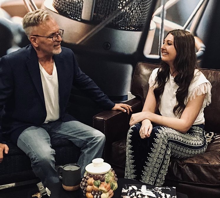 Two women sitting on a sofa, smiling and talking, with a jar of candies and a coffee cup on a table in front of them.