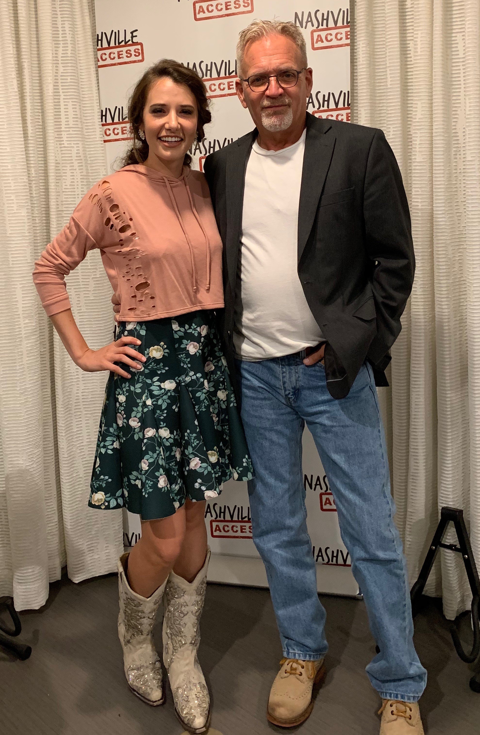 A woman and a man stand together in front of a Nashville Access backdrop, smiling. The woman wears a pink top, floral skirt, and cowboy boots. The man wears a black blazer, white shirt, and jeans.