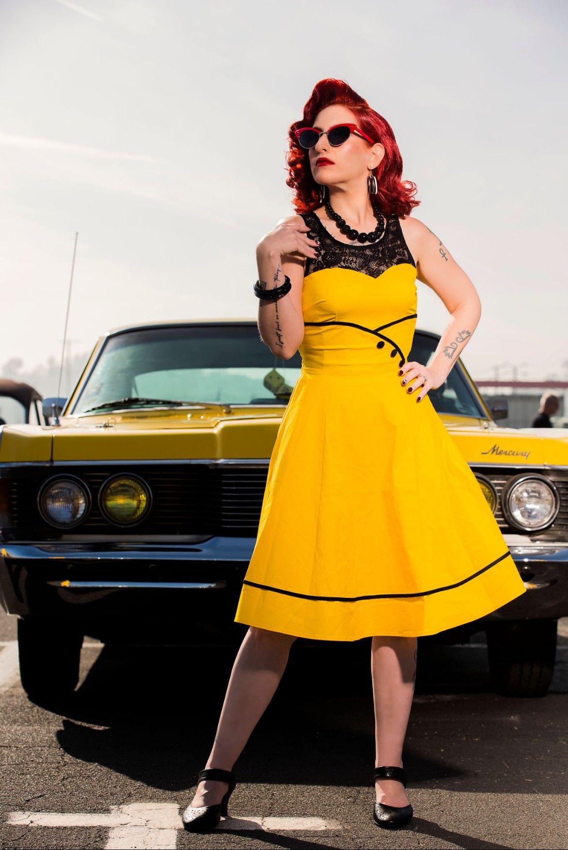 Woman with red hair in a yellow dress and sunglasses stands in front of a vintage yellow car outdoors.