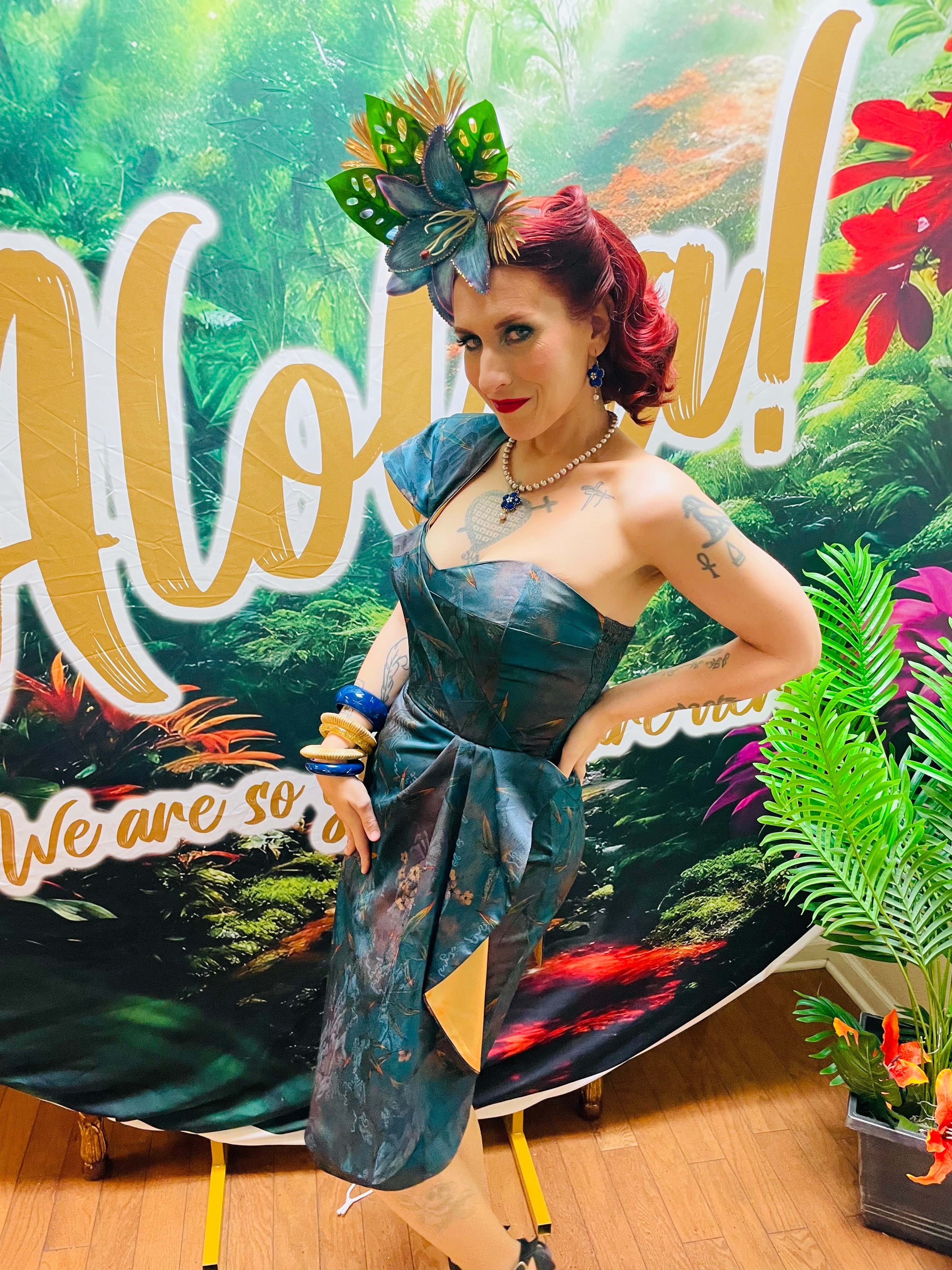 Woman with red hair and butterfly headpiece poses in front of a tropical-themed backdrop, wearing a strapless dress and jewelry.